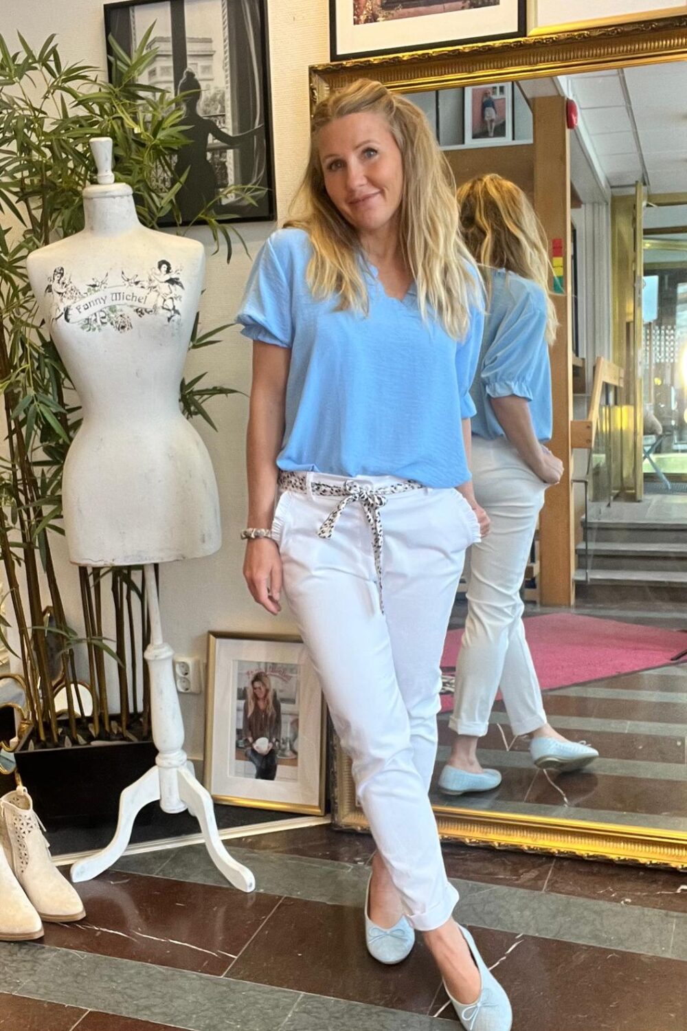 Kvinna i ljusblus och vita byxor poserar och ler framför en stor guldspegel i en butiksmiljö. Bredvid står en provdocka med texten “Fanny Michel”, omgiven av växter, tavlor och skor på golvet.