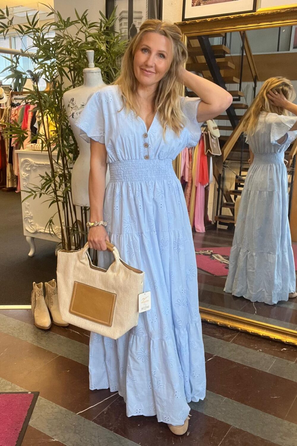 Kvinna i ljusblå lång klänning med broderie anglaise poserar i en klädbutik, håller en stor beige handväska och har ena handen i håret. Spegel bakom visar baksidan; klädställningar och skor syns.