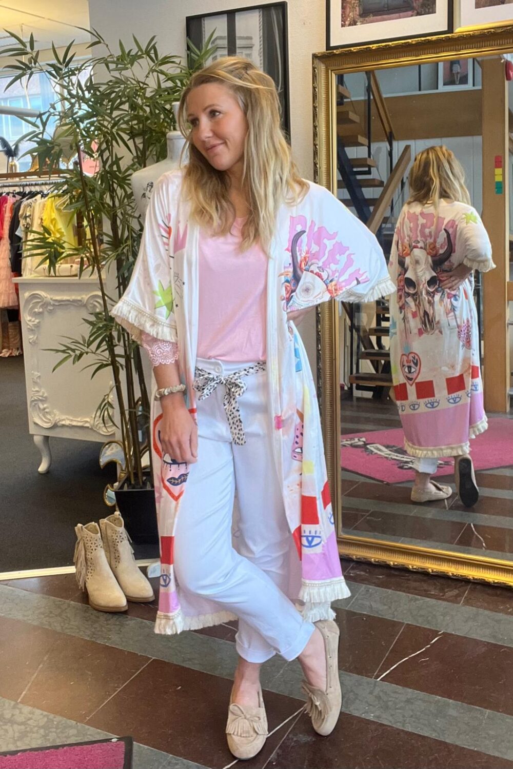 Kvinna står avslappnat i en klädbutik och poserar med benen i kors, iklädd rosa topp, vita byxor och en lång mönstrad kimono. Stor guldspegel visar ryggen med djurkraniemotiv; klädställning och växt i bakgrunden.