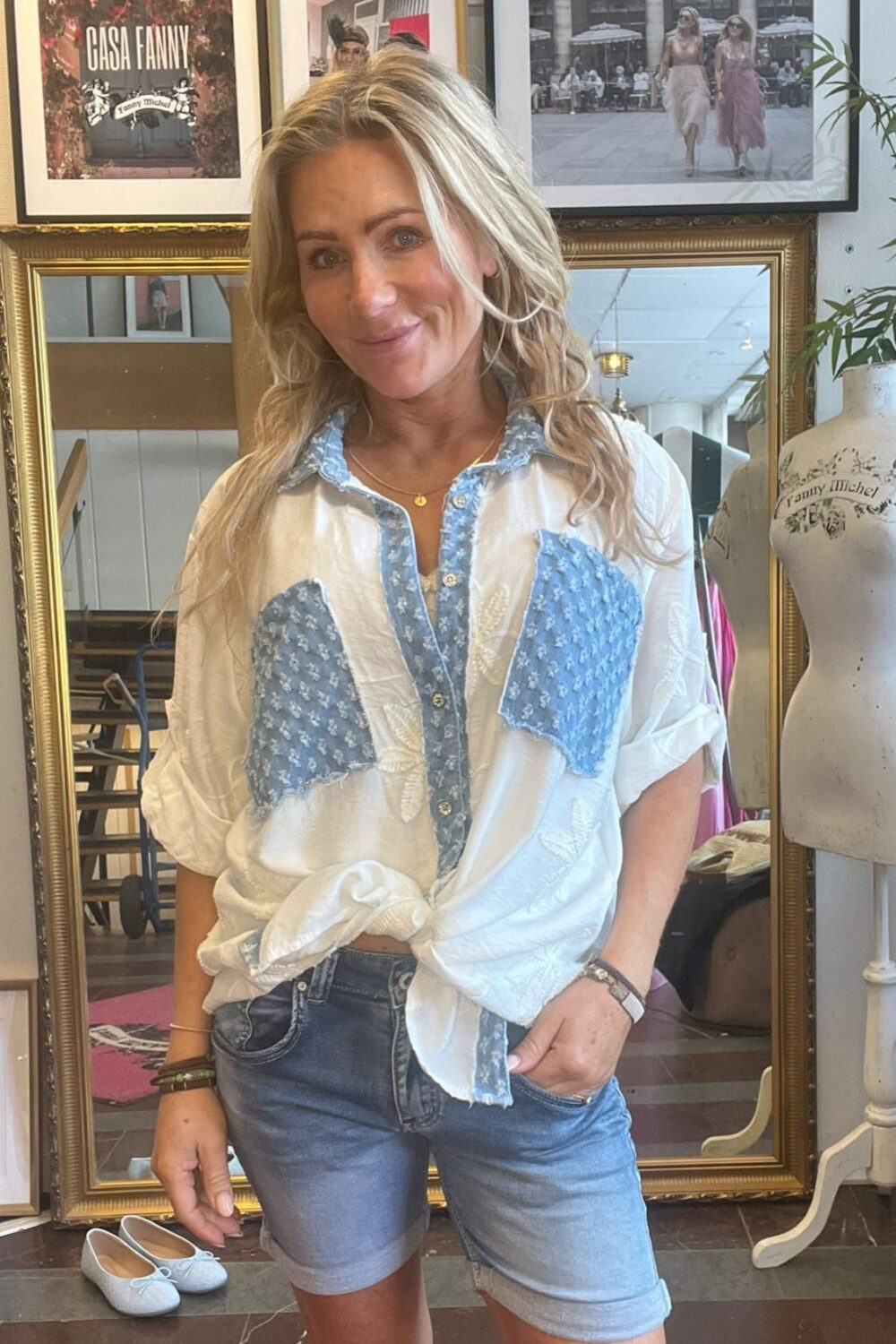 Leende kvinna står framför en stor guldramsspegel och visar en vit blus med blå mönstrade partier och jeansshorts, handen i fickan. I bakgrunden syns posters med texten “CASA FANNY” och en provdocka.