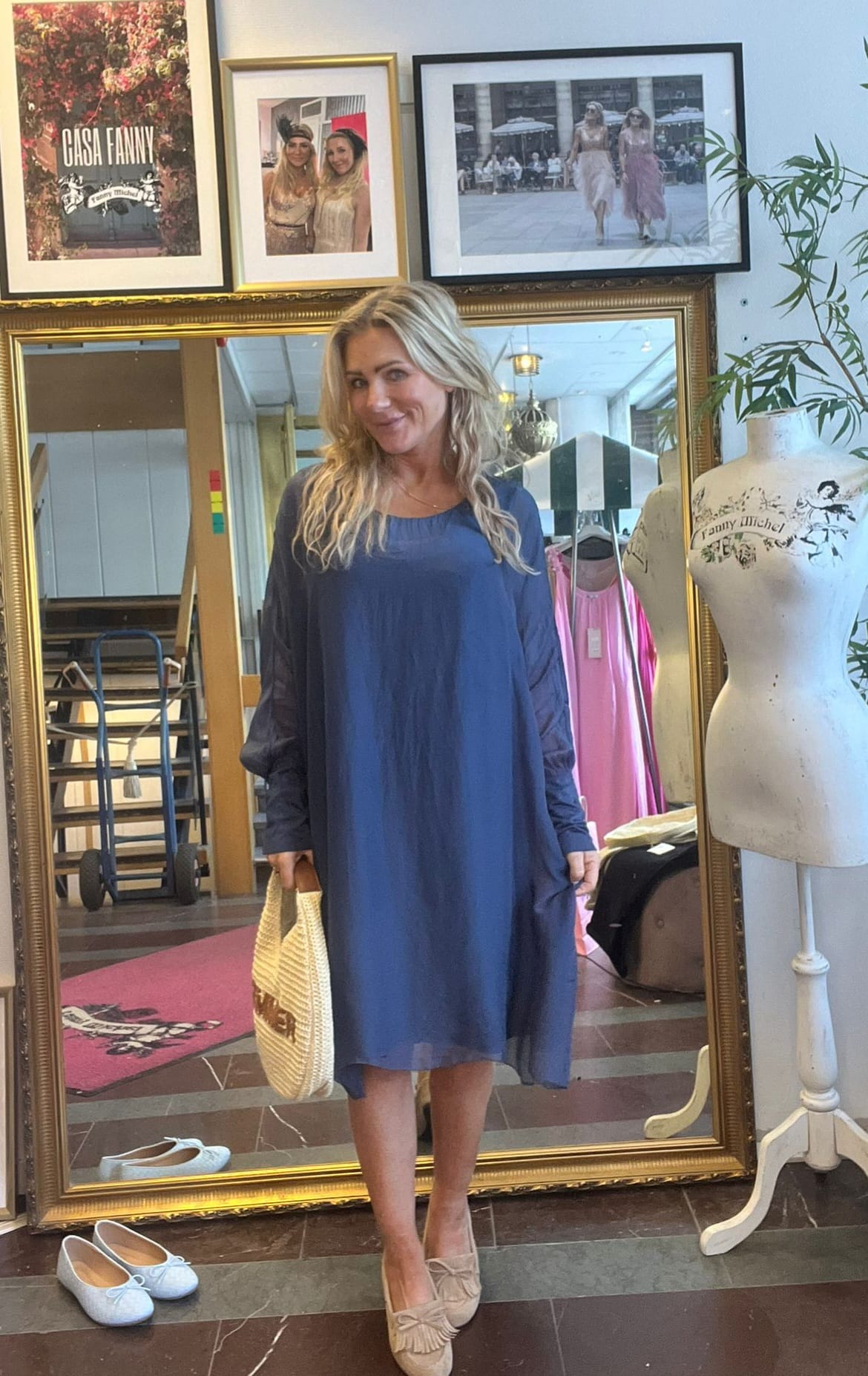 Blond kvinna står och ler framför en stor guldramsspegel i en butik, klädd i blå, lös klänning och håller en stråväska. På väggen syns inramade foton och texten ”CASA FANNY” samt provdocka.