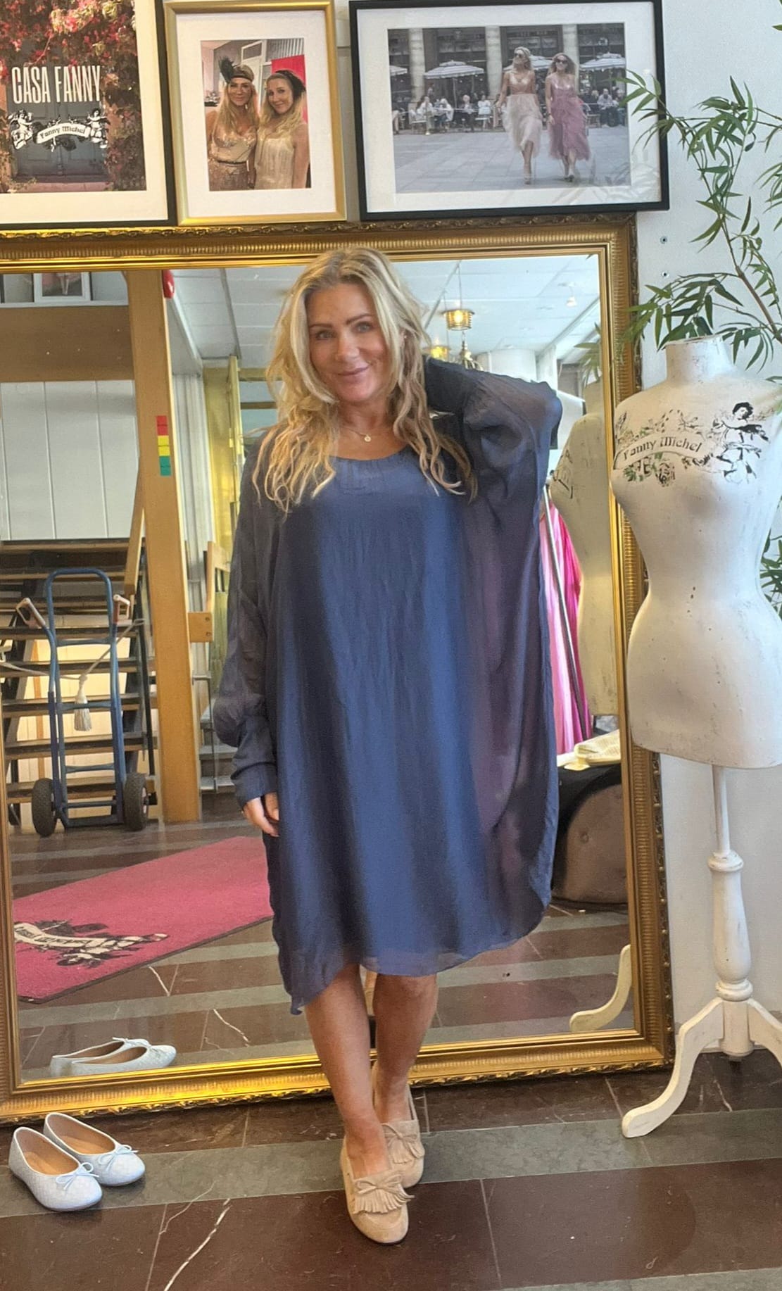 Blond kvinna står och ler framför en stor guldramsspegel i en ateljé eller butik, iklädd blå klänning och tunn mörk kimono. Bredvid syns provdocka med texten ”Fanny Michel” och inramade foton.