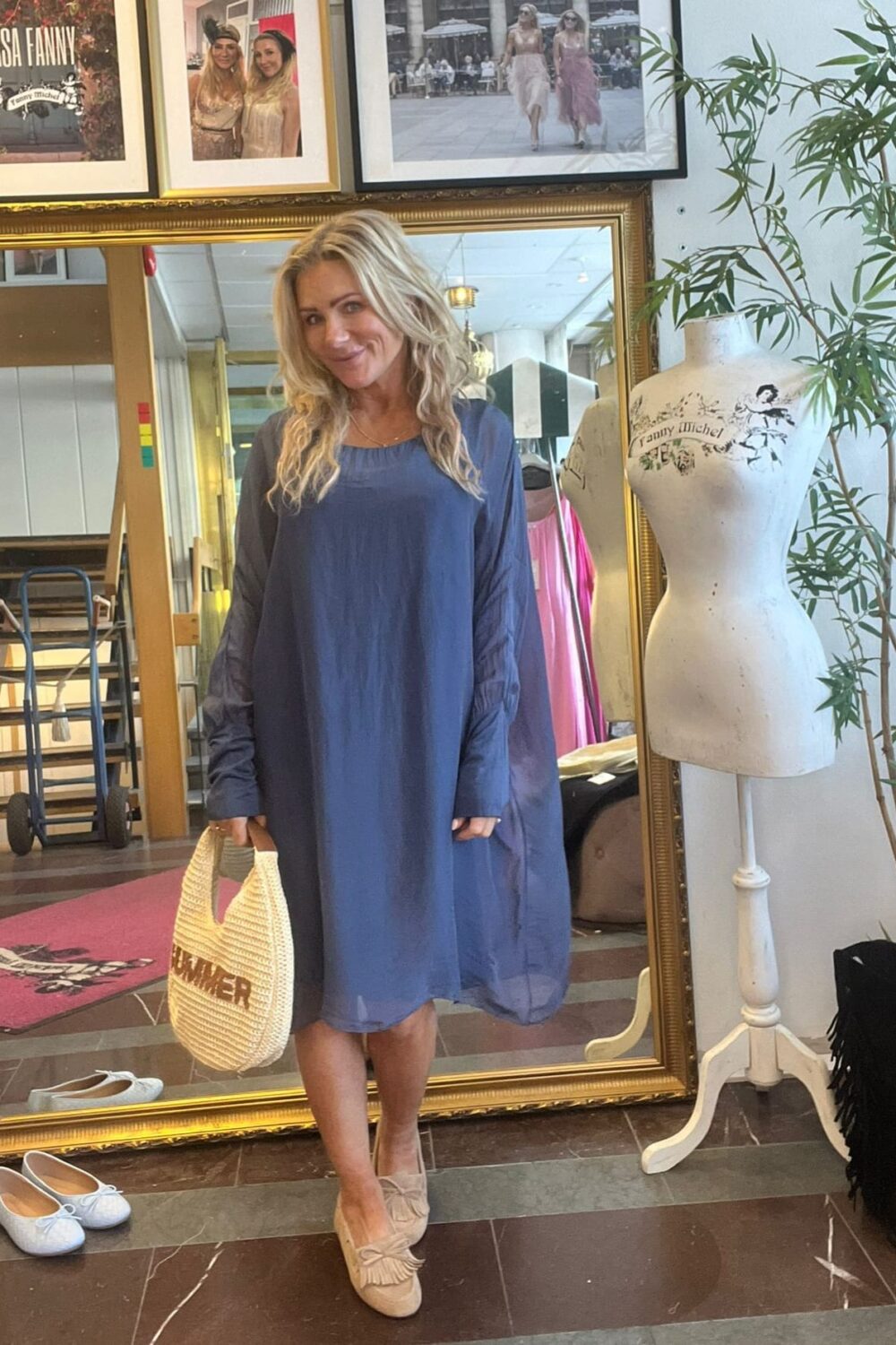 Leende kvinna i blå, långärmad klänning står framför en stor guldramsspegel och håller en beige vävd väska med texten ”SUMMER”. Intill syns en provdocka med text ”Fanny Michel”, skor på golvet och inramade foton.