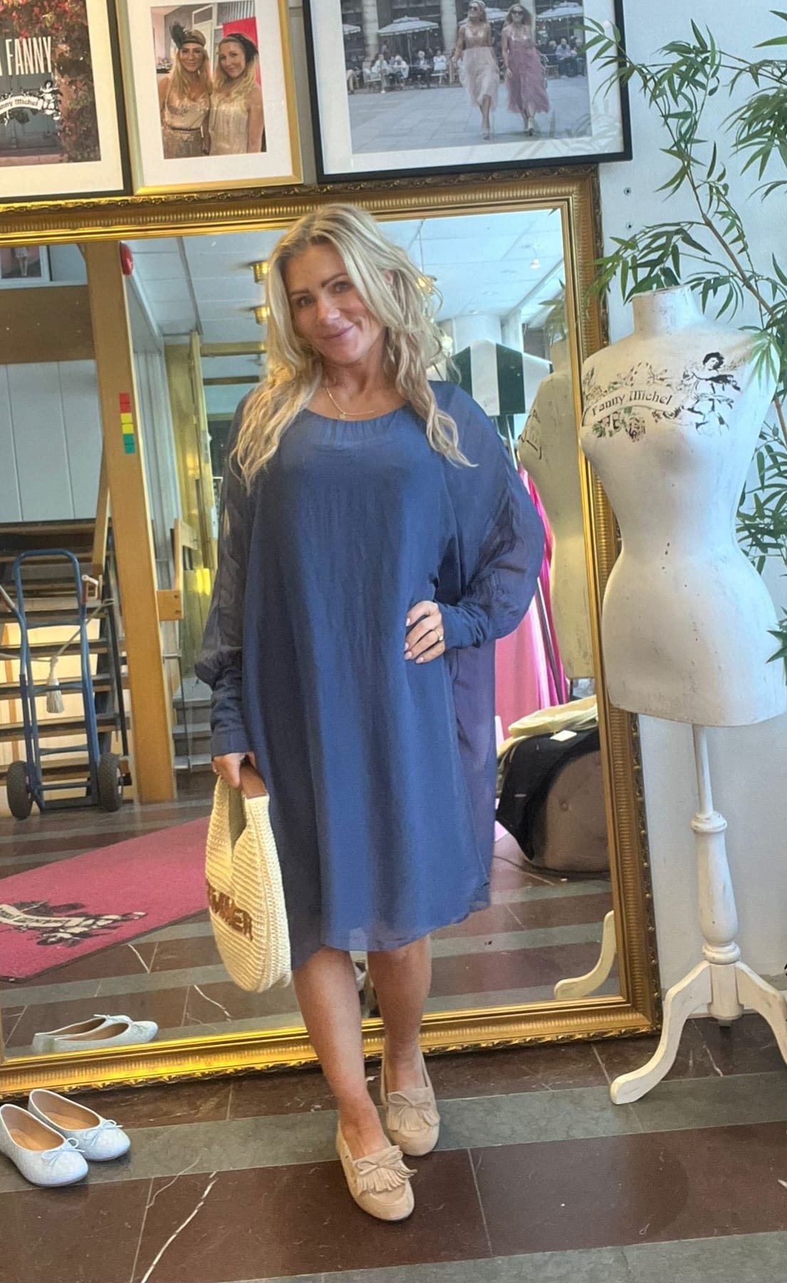 Blond kvinna står och ler framför en stor guldramsspegel, klädd i en lös blå klänning och beige skor, med en stråväska i handen. Bredvid syns en provdocka med texten ”Fanny Michel” och inramade foton.