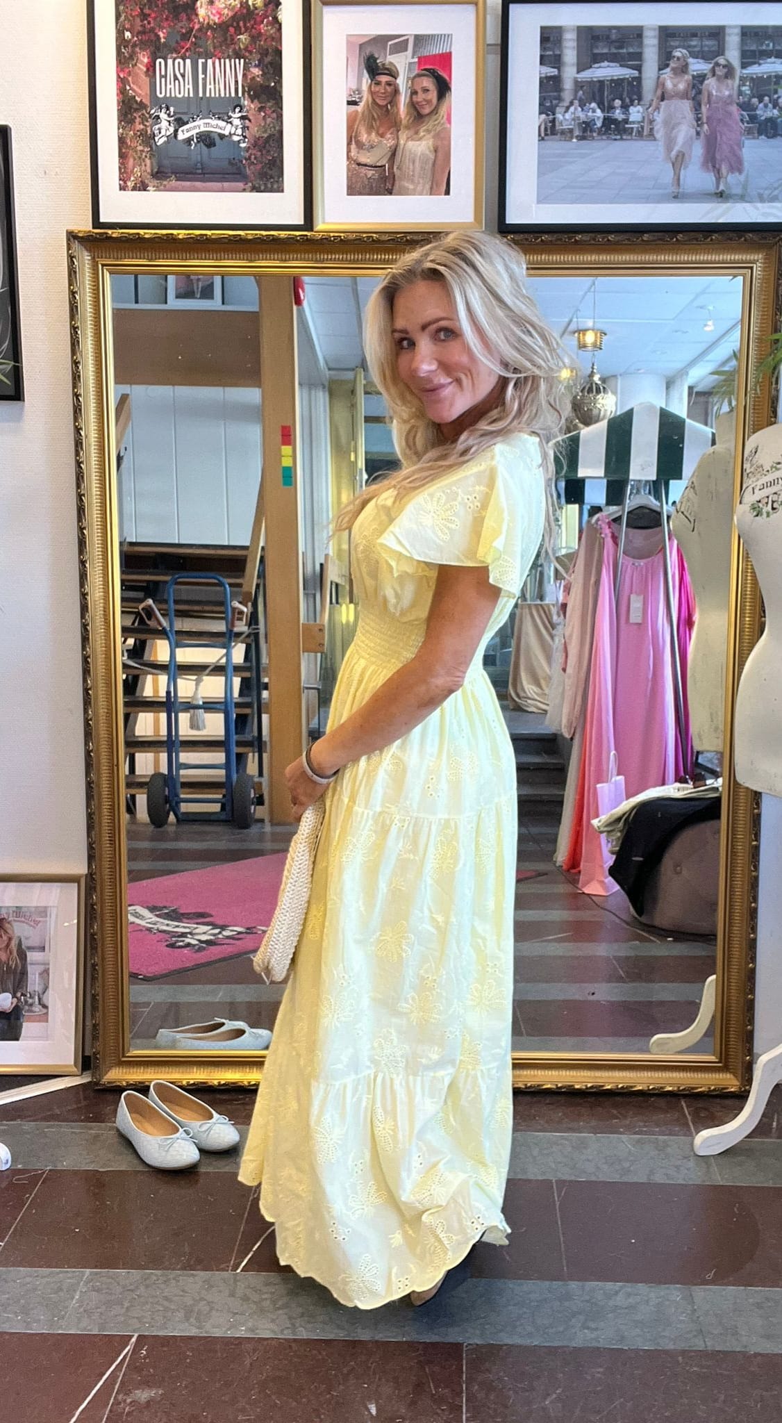 Kvinna i ljusgul, broderad långklänning poserar och ler i en stor guldramsspegel i en butik. Hon håller en vit väska; skor ligger på golvet och kläder hänger bakom. Text på tavla: “CASA FANNY”.