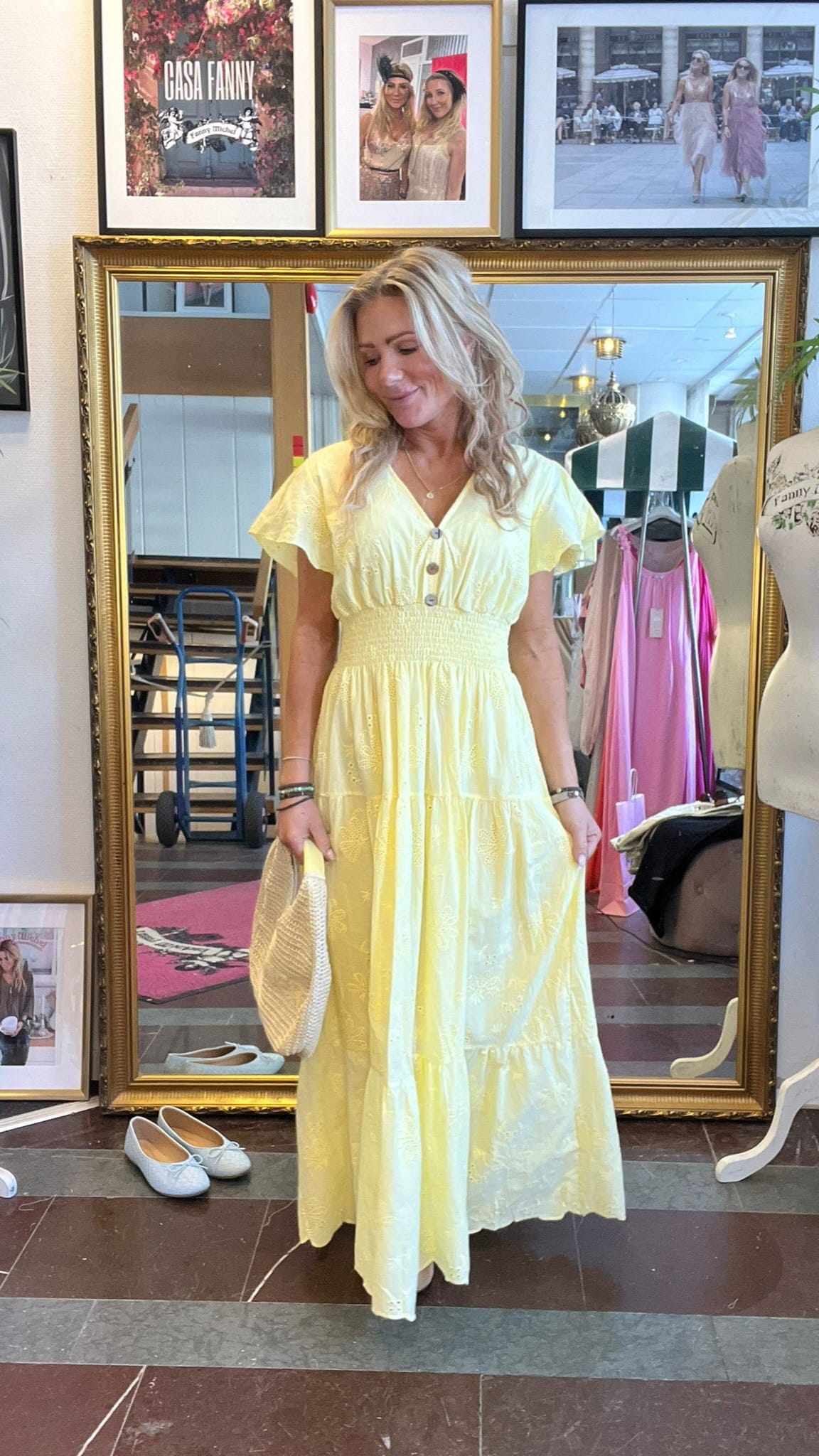 Blond kvinna står framför en stor guldramsspegel och tittar ned med ett leende, klädd i ljusgul lång klänning och håller en stråväska. I bakgrunden syns butiksmiljö, skor på golvet och tavlor med texten “CASA FANNY”.