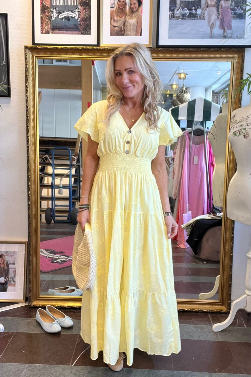 Kvinna står och poserar framför en stor guldramad spegel i en klädbutik, iklädd ljusgul lång klänning med knappar och volanger och håller en stråväska. På väggen syns inramade foton och texten “CASA FANNY”.