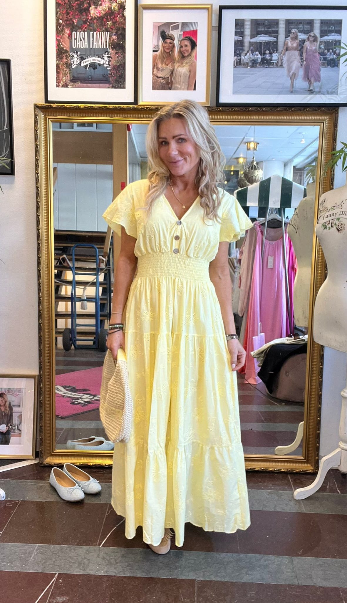 Kvinna står och poserar framför en stor guldramad spegel i en klädbutik, iklädd ljusgul lång klänning med knappar och volanger och håller en stråväska. På väggen syns inramade foton och texten “CASA FANNY”.