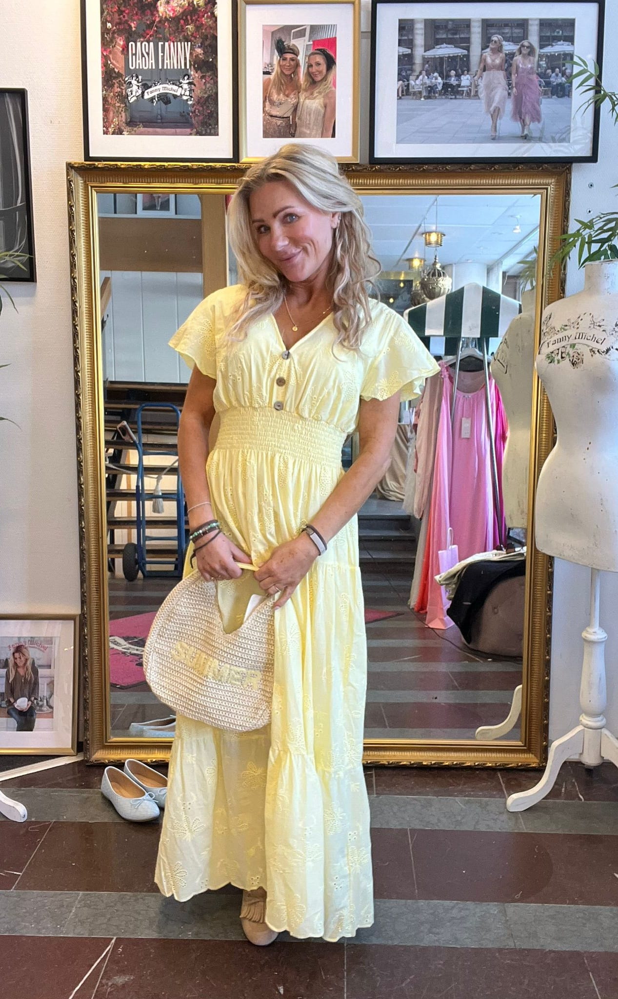 Kvinna poserar framför en stor guldramsspegel i en butik, klädd i ljusgul långklänning och håller en flätad väska. Runtom syns skyltdocka och rosa klänning. Inramad text: ”CASA FANNY”.
