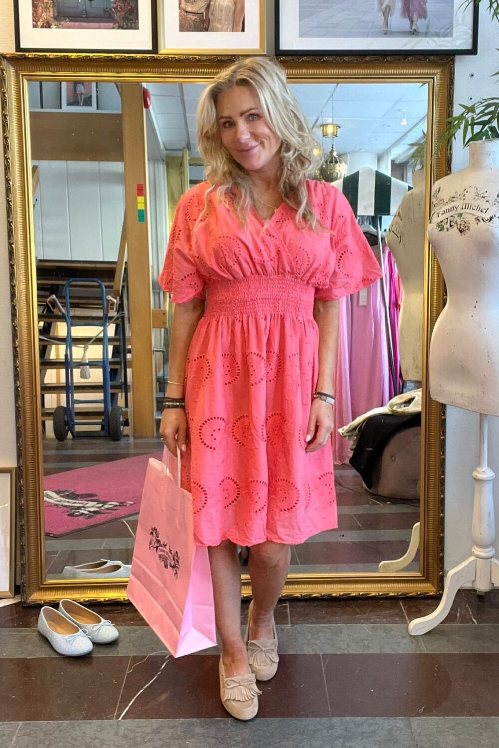 Blond kvinna står och poserar framför en stor guldramad spegel i en butik, iklädd korallrosa klänning och beige loafers, och håller en rosa shoppingpåse. På väggen syns inramade foton med texten ”CASA FANNY” och ”Fanny Michel”.
