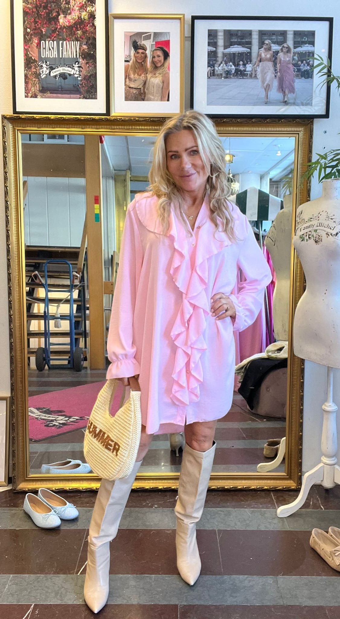 Blond kvinna poserar framför en stor guldramsspegel i en studio/butik, klädd i ljusrosa volangklänning och vita knähöga stövlar, med stråväska. På väggen hänger inramade foton med texten ”CASA FANNY” och ”Fanny Michel”.
