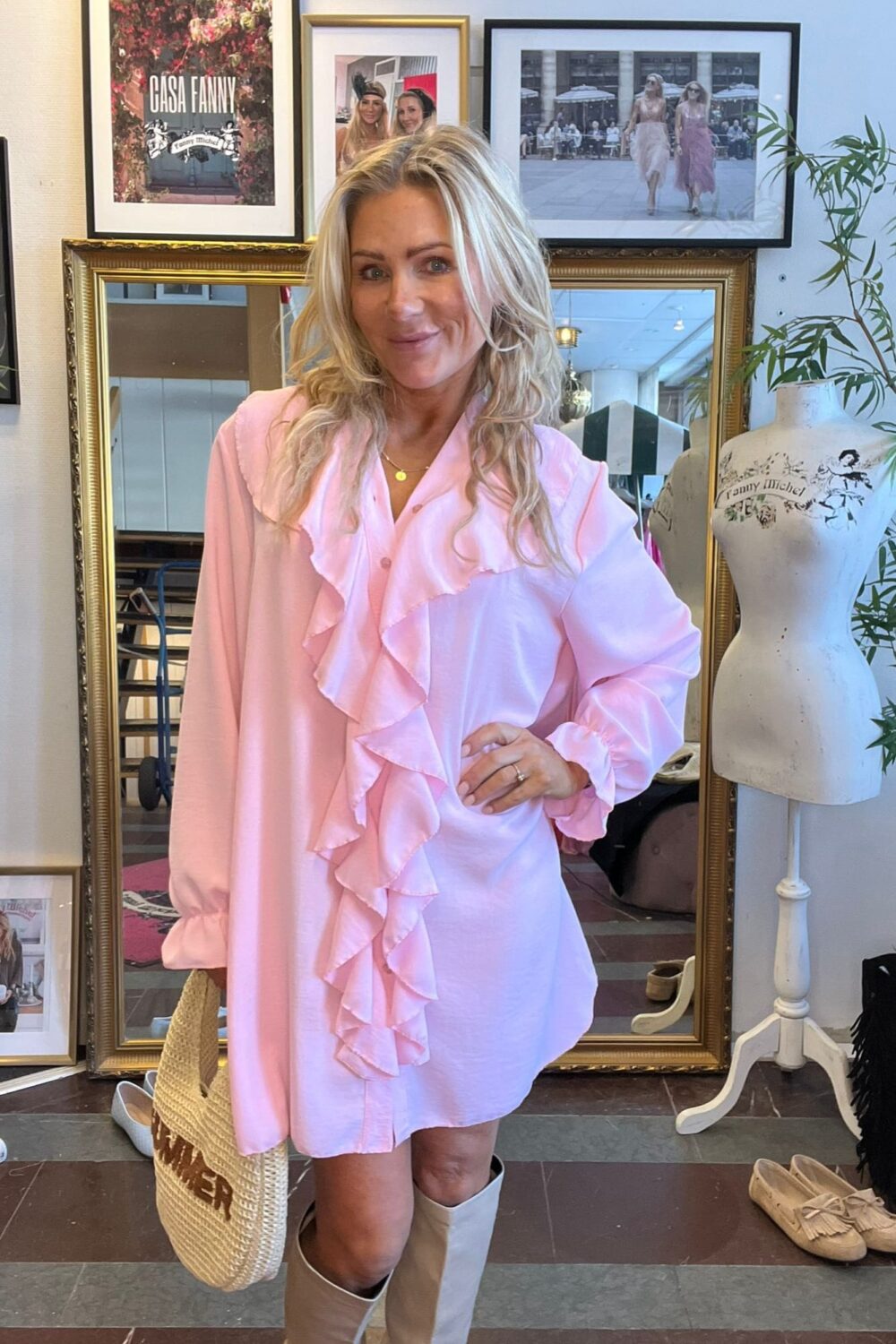 Blond kvinna poserar och ler i en butik, klädd i ljusrosa volangklänning och ljusa stövlar, med stråväska i handen. Bakom syns inramade foton med texten “GÅSA FANNY” och en provdocka märkt “Fanny Michel”.