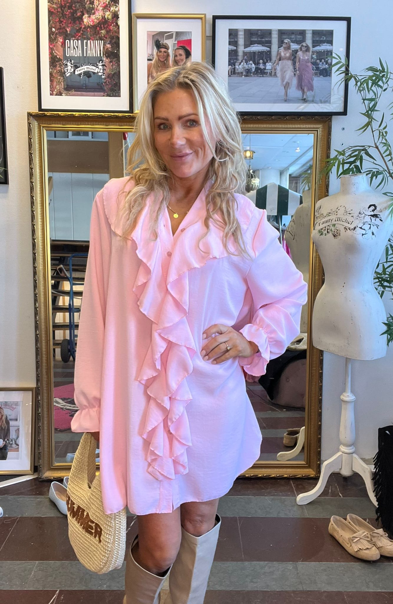 Blond kvinna poserar och ler i en ljusrosa volangklänning, håller en halmväska och bär ljusa stövlar. Hon står i en butik med stor spegel, klädskor och ramar; texten “GÅSA FANNY” syns.