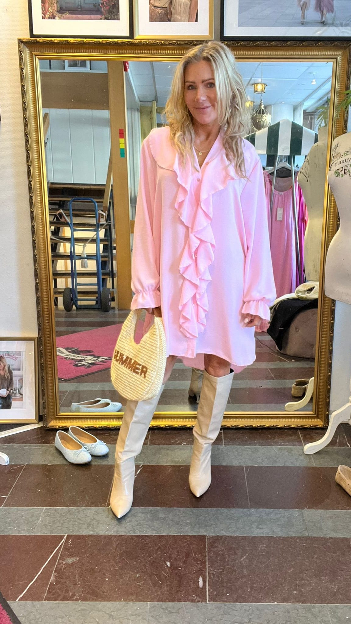 Kvinna poserar framför en stor guldramsspegel i en butik, klädd i ljusrosa volangklänning och vita knähöga stövlar. Hon håller en halmväskan med texten ”SUMMER”; skor och kläder syns runtom.