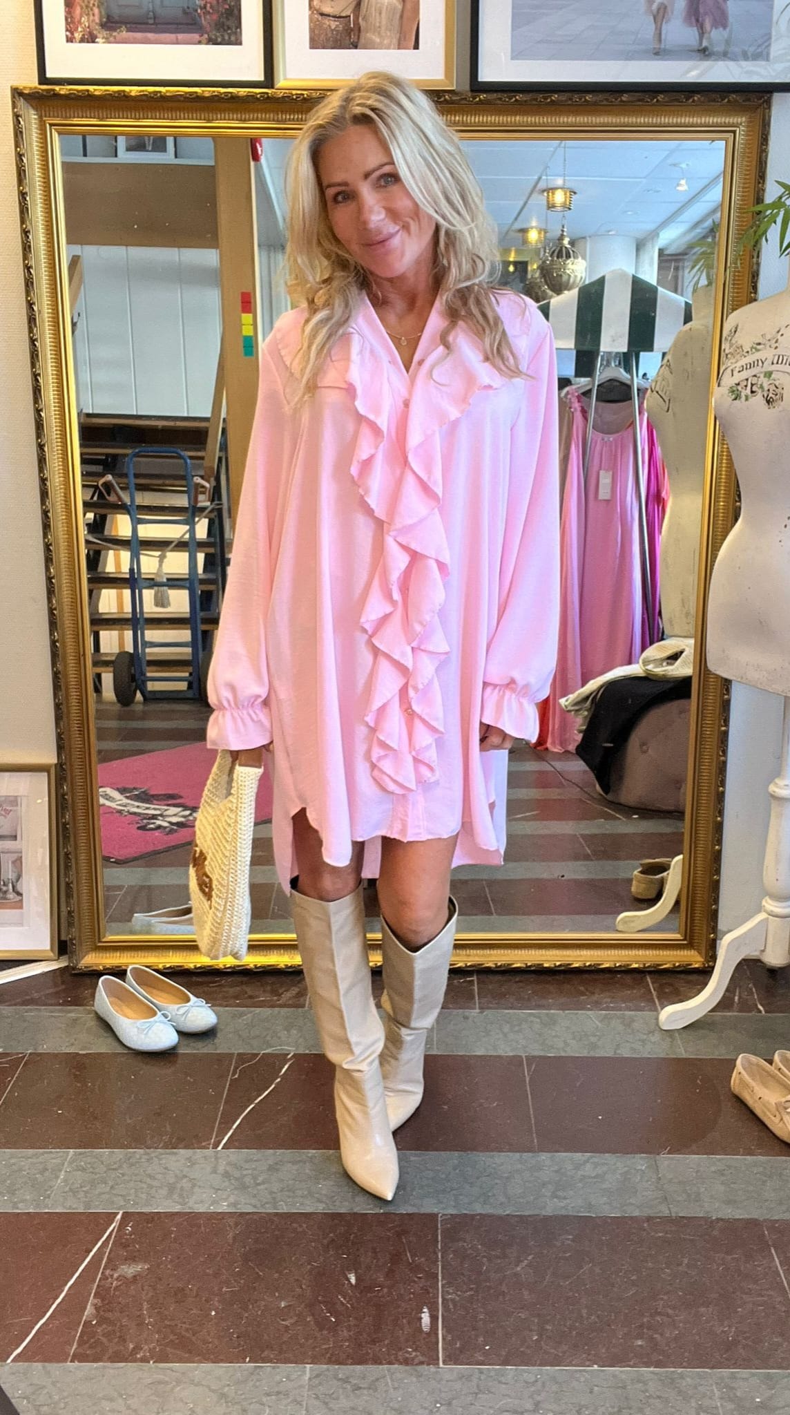 Blond kvinna poserar framför en stor guldramsspegel i en klädbutik, iklädd en ljusrosa volangklänning och vita knähöga stövlar. Hon håller en flätad väska; runtom syns klädställning, provdocka och skor på golvet.
