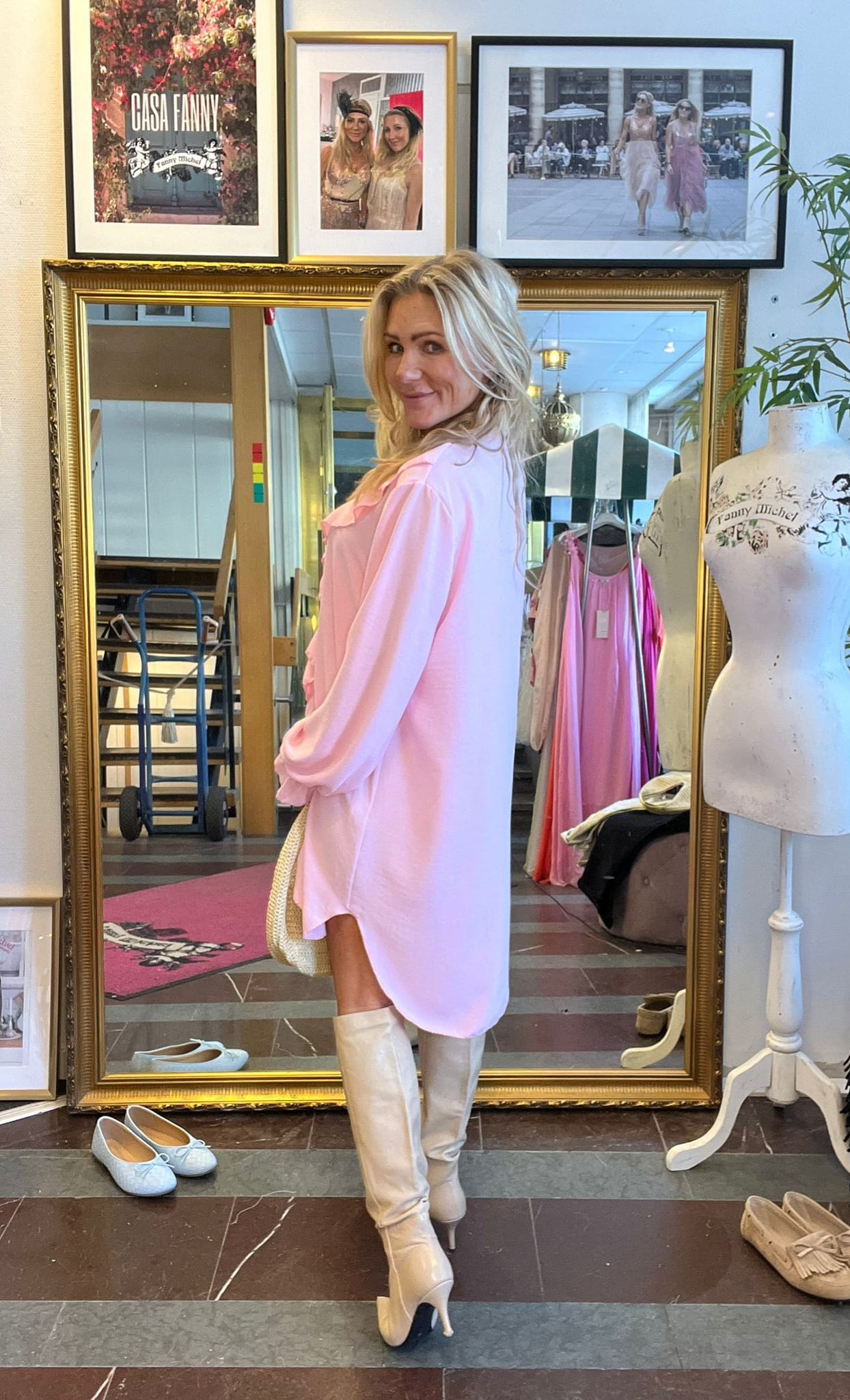 Blond kvinna poserar i profil framför en stor guldramsspegel, klädd i ljusrosa klänning och vita högklackade stövlar. Runtom syns en butiksmiljö med kläder, skyltdocka och inramade foton med texten “GÅSA FANNY”.