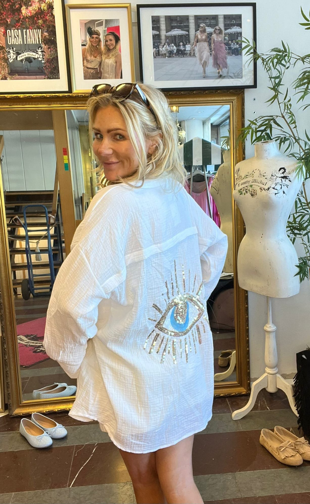 Leende kvinna vänder sig mot kameran och visar baksidan av en vit skjortklänning med paljettbroderat öga. Hon står i en ateljé/butik med spegel, provdocka med texten “Fanny Michel” och inramade foton.