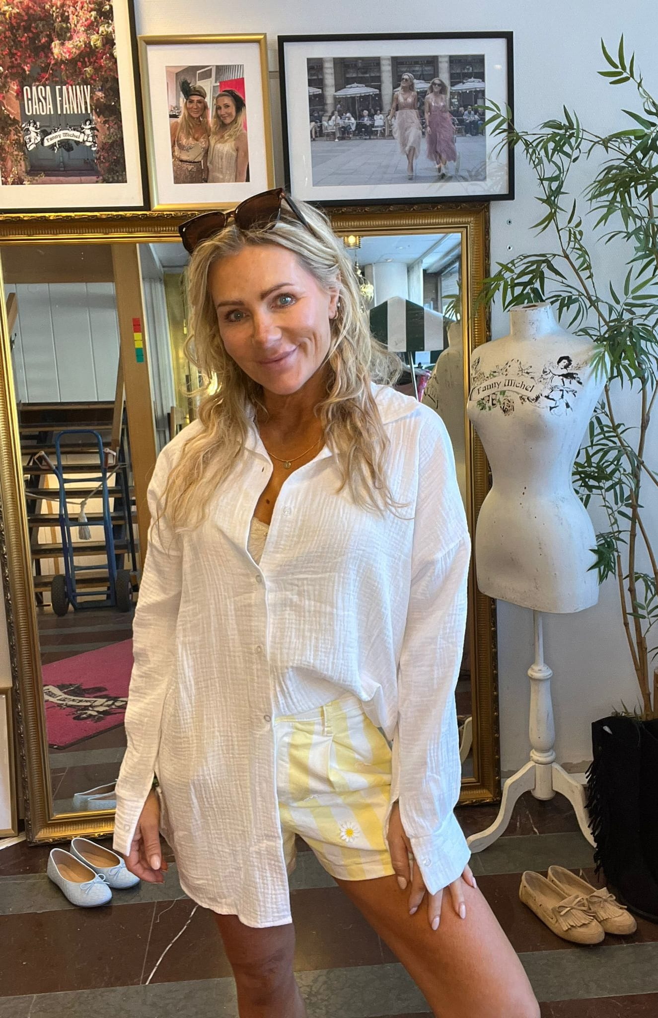 Blond kvinna i vit skjorta och gulrutiga shorts med solglasögon på huvudet ler mot kameran i en butik/ateljé. Bakom henne syns inramade foton och texten “CASA FANNY” samt provdocka märkt “Fanny Michel”.