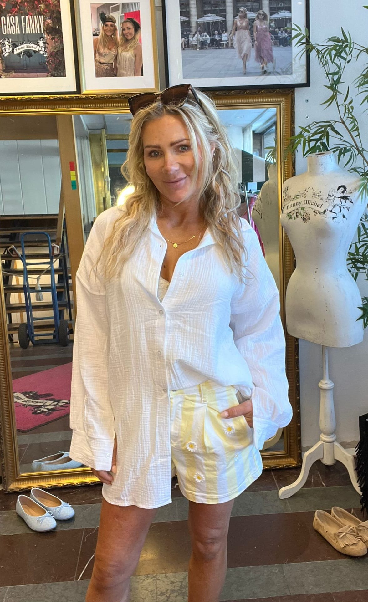 Leende kvinna med solglasögon på huvudet står och poserar i en butik eller ateljé, iklädd vit skjorta och gulvit randig kjol. Bakom syns inramade foton med texten “CASA FANNY” och en provdocka märkt “Fanny Michel”.