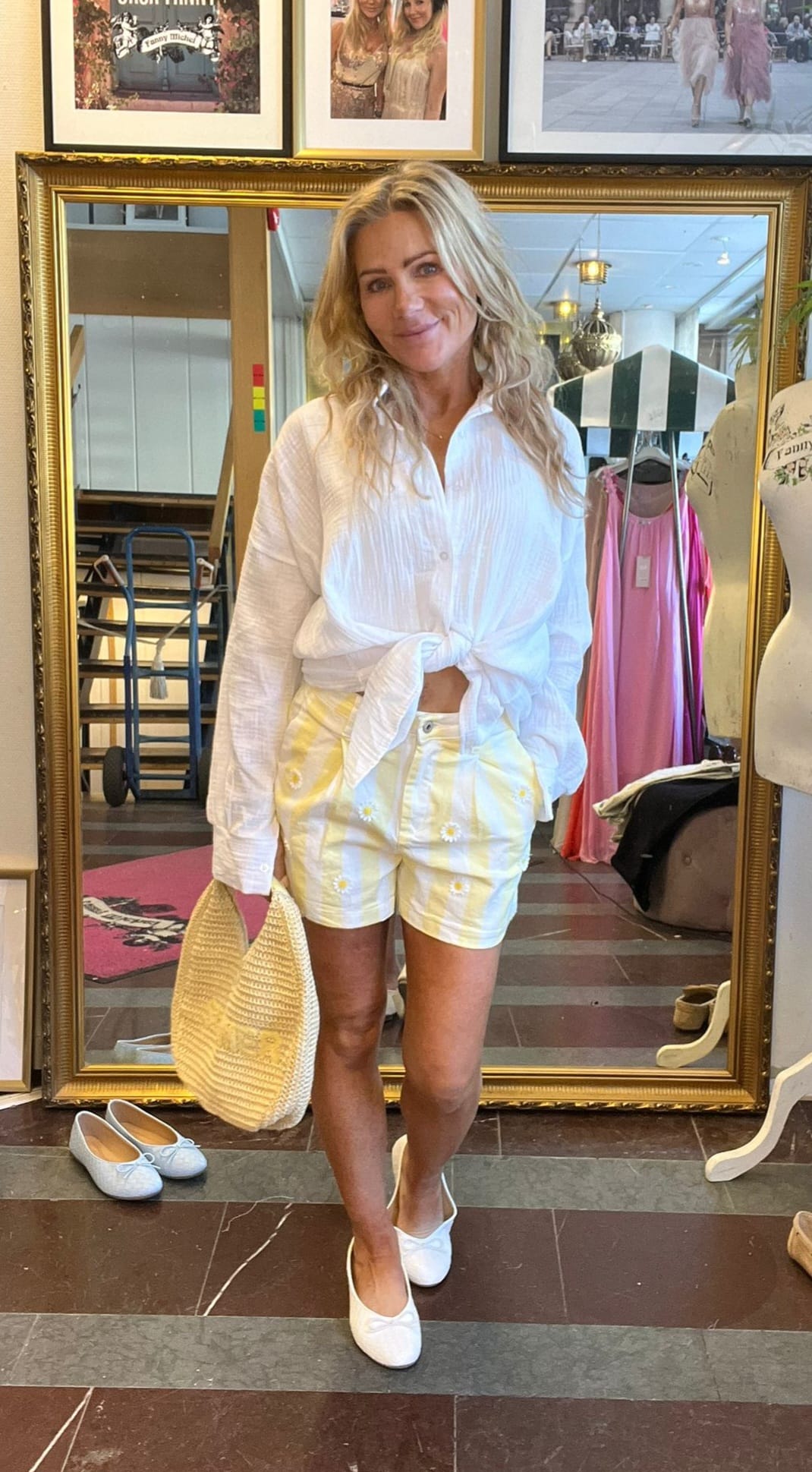Leende kvinna poserar framför en stor guldramsspegel i en butik, klädd i vit knuten skjorta och gulvit randiga shorts med små blommor, håller en stråväska och bär vita ballerinaskor.