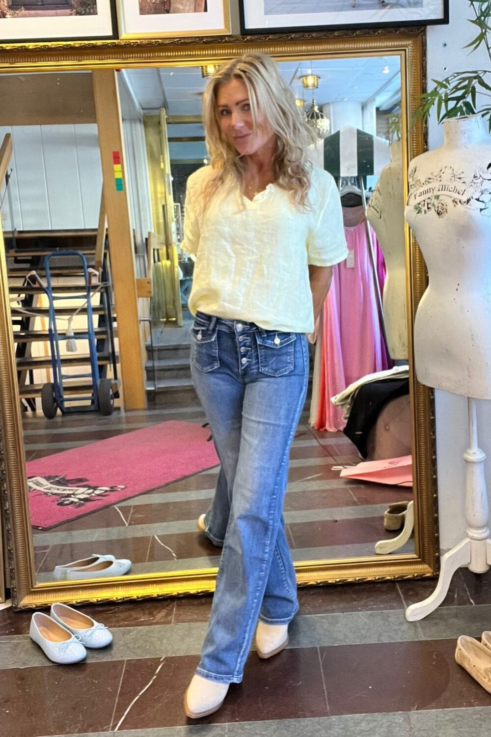 Blond kvinna poserar framför en stor guldramsspegel i en butik eller ateljé, klädd i vit blus, blå jeans och vita skor. I bakgrunden syns provdocka med texten “Funny Mickey”, klänningar och inramade foton.