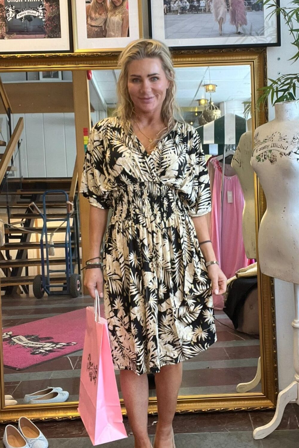 Leende kvinna står i en butik och håller en rosa shoppingpåse, klädd i svartvit blommig klänning och beige skor. Bakom syns stor guldramsspegel, skyltdocka och inramade foton med texten “CASA FANNY”.