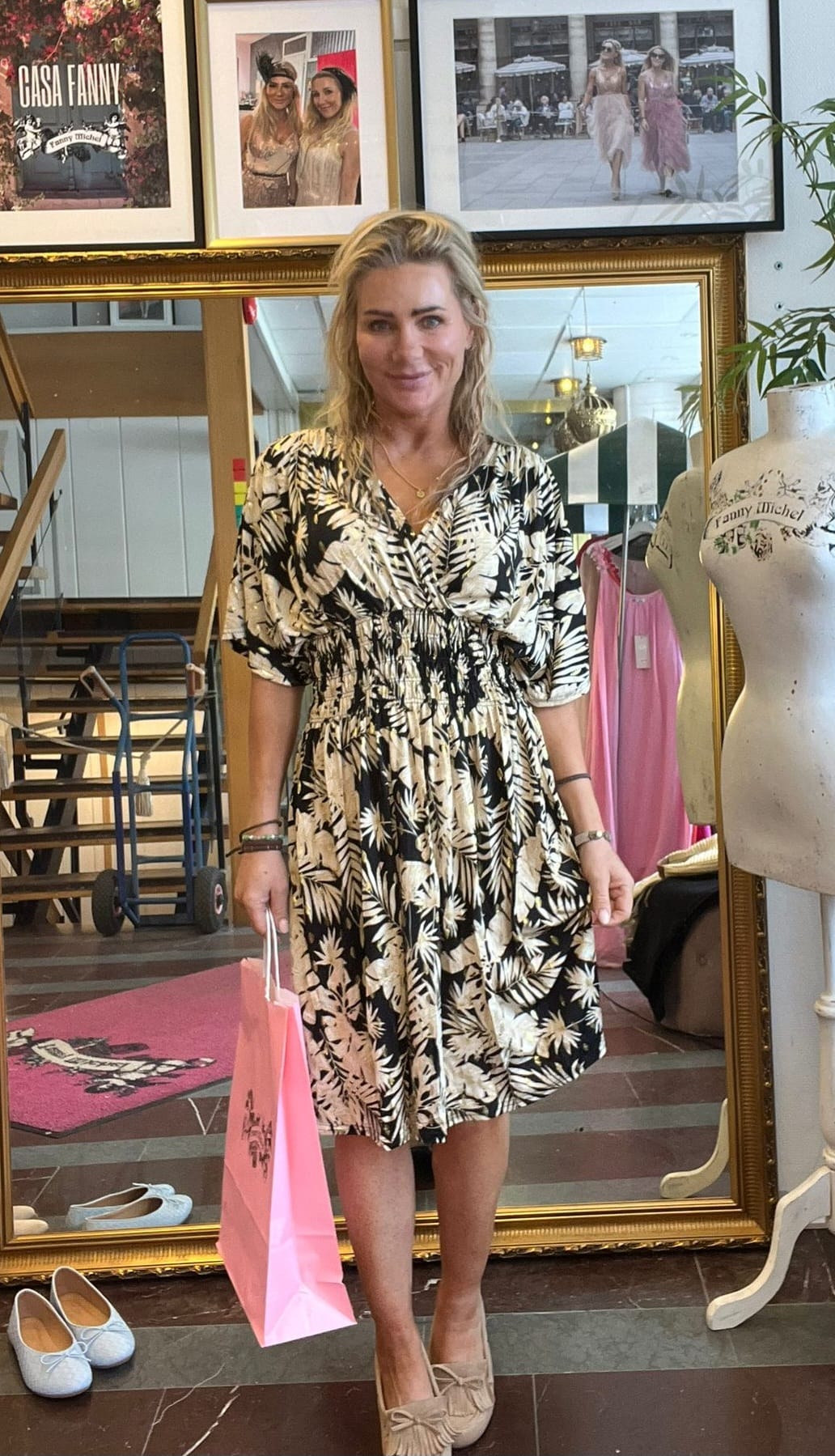 Leende kvinna står i en butik och håller en rosa shoppingpåse, klädd i svartvit blommig klänning och beige skor. Bakom syns stor guldramsspegel, skyltdocka och inramade foton med texten “CASA FANNY”.