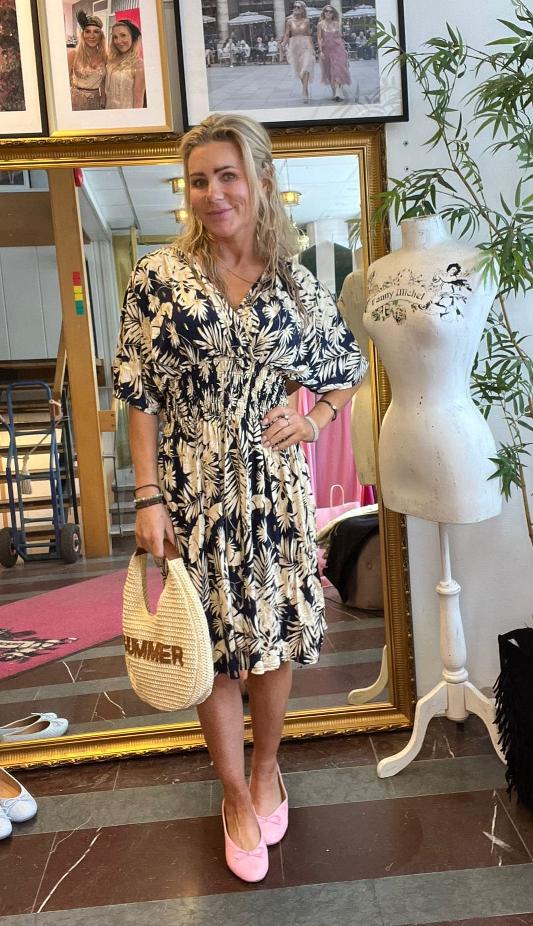 Blond kvinna poserar med handen på höften framför en stor guldramsspegel i en butik eller ateljé, bär blommig klänning och rosa skor samt håller en stråväska med texten “SUMMER”; intill står en provdocka märkt “Fanny Michel”.