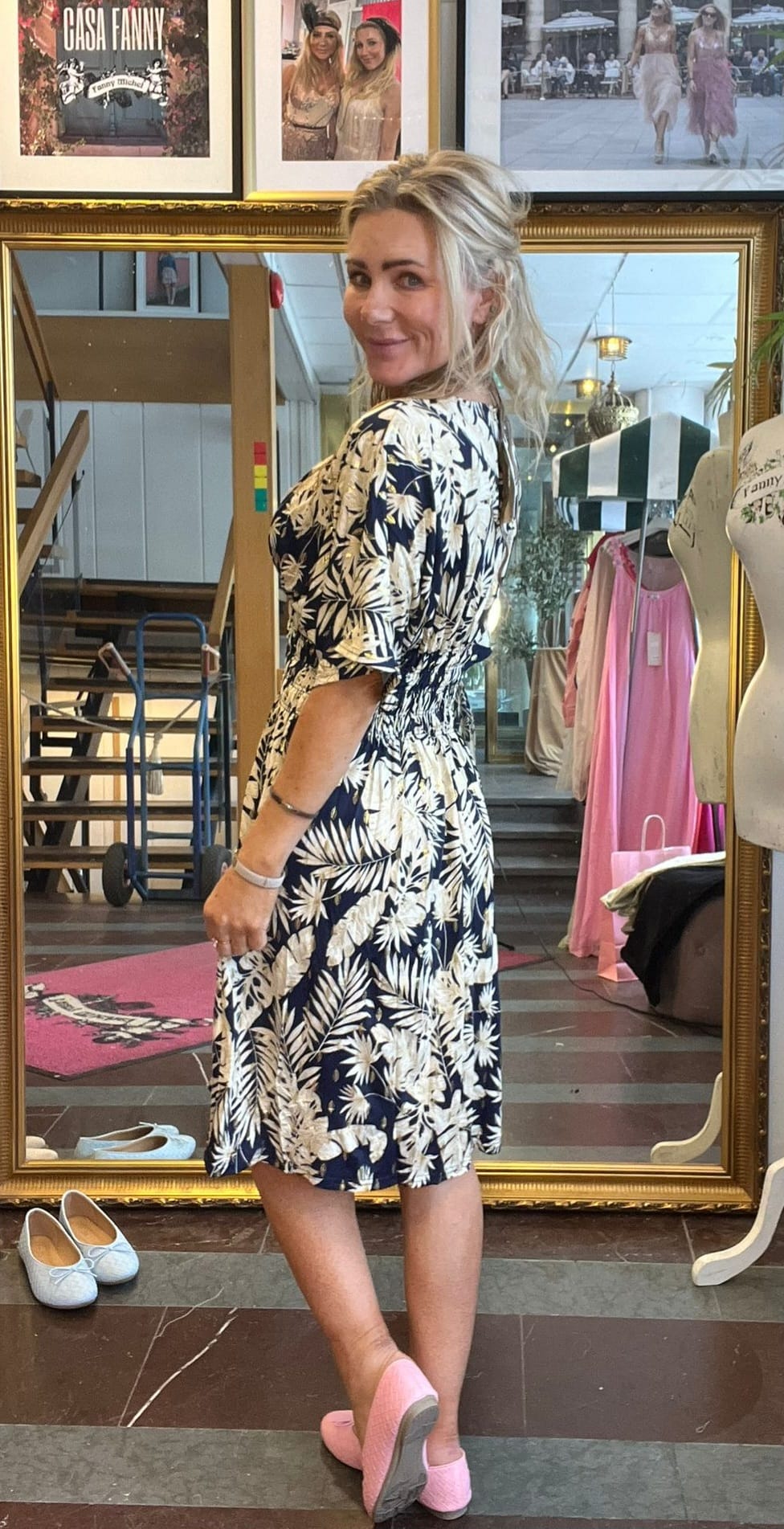 Blond kvinna i mörkblå klänning med vitt bladmönster poserar framför en stor guldramsspegel och tittar över axeln, i en butiksmiljö med klädställning och skor på golvet. Inramad väggbild med texten ”CASA FANNY”.