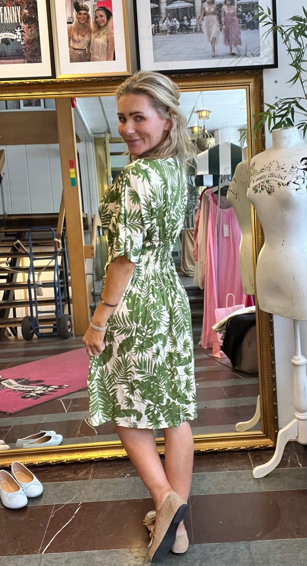 Kvinna står framför en stor spegel och provar en grönvit klänning med bladmönster, vänd halvt om och ler mot kameran. I bakgrunden syns provrum, rosa klänning och skyltdocka med texten “Fanny Michel”.