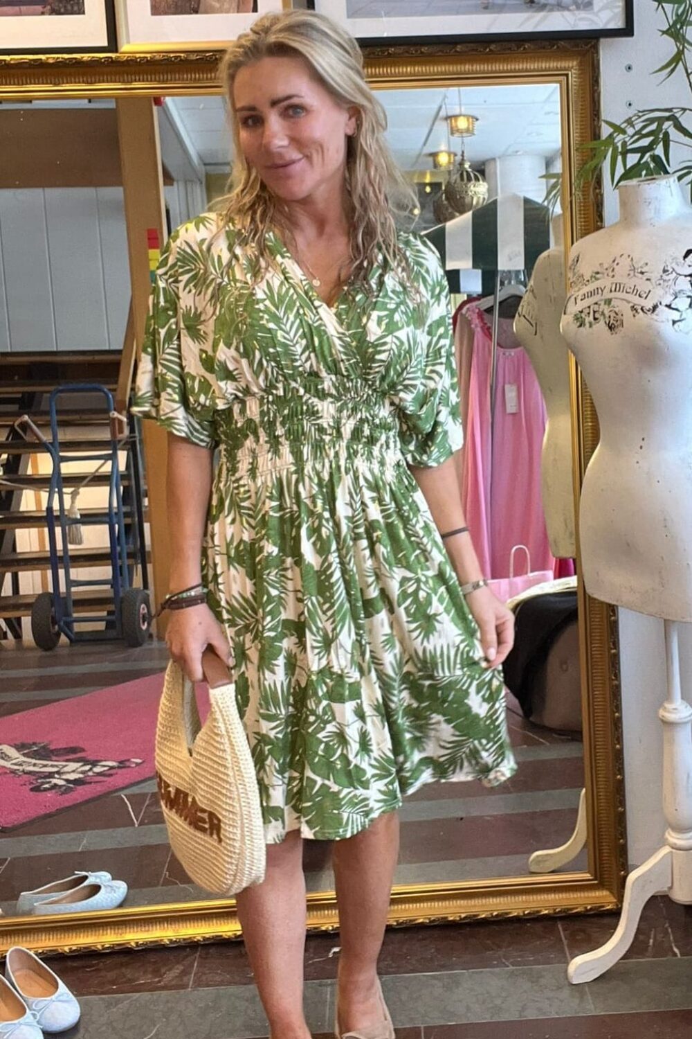Kvinna står och posar i en butiksmiljö, klädd i grönvit bladmönstrad klänning och ljusa loafers, med en stråväska i handen. Bakom syns en stor spegel, modefoton och en provdocka med texten “Fanny Michel”.