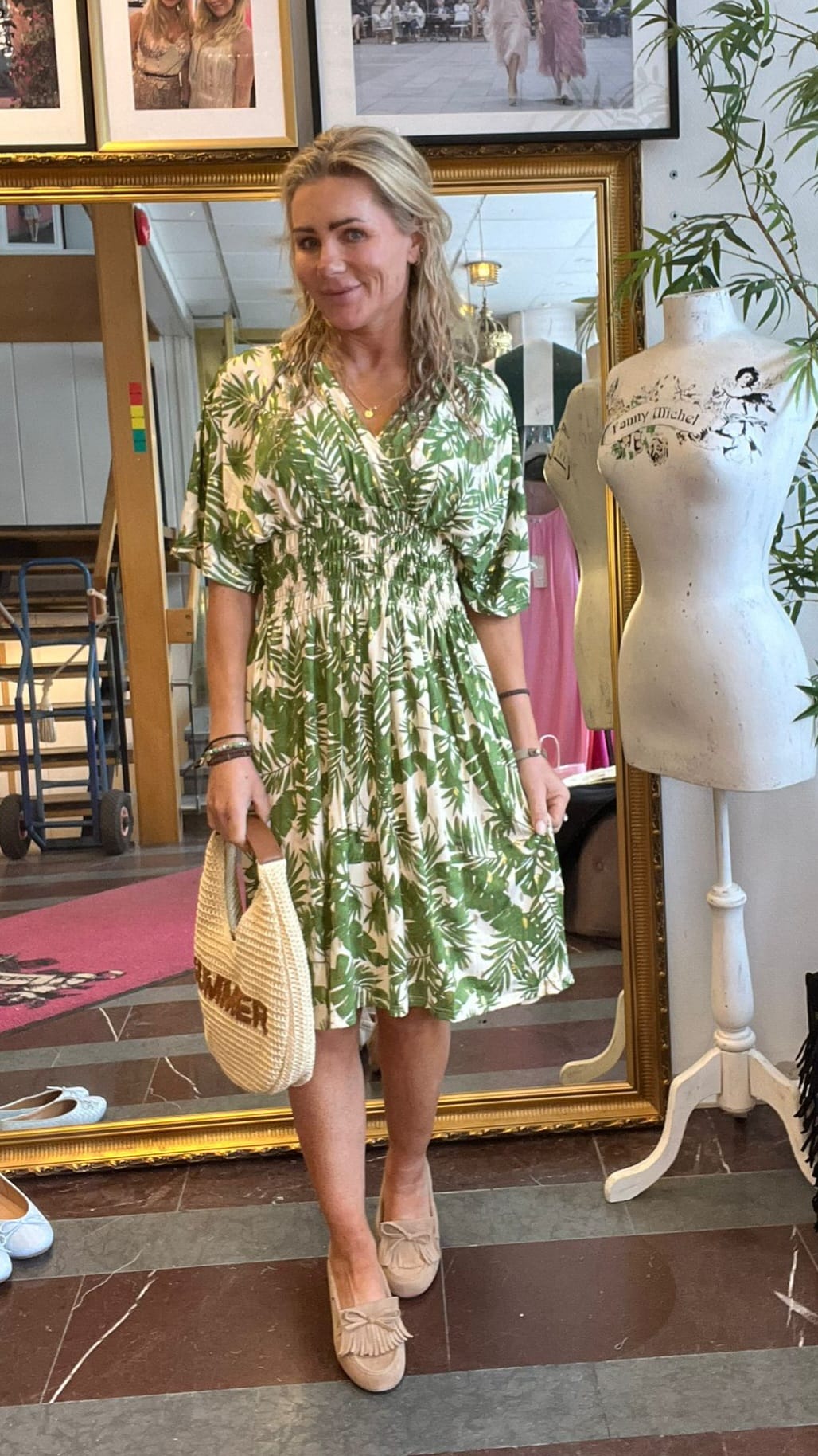 Leende kvinna står framför en stor guldramsspegel i en butik, klädd i grönvit bladmönstrad klänning och beige loafers, och håller en flätad väska. Bredvid står en provdocka med texten “Fanny Michel”.