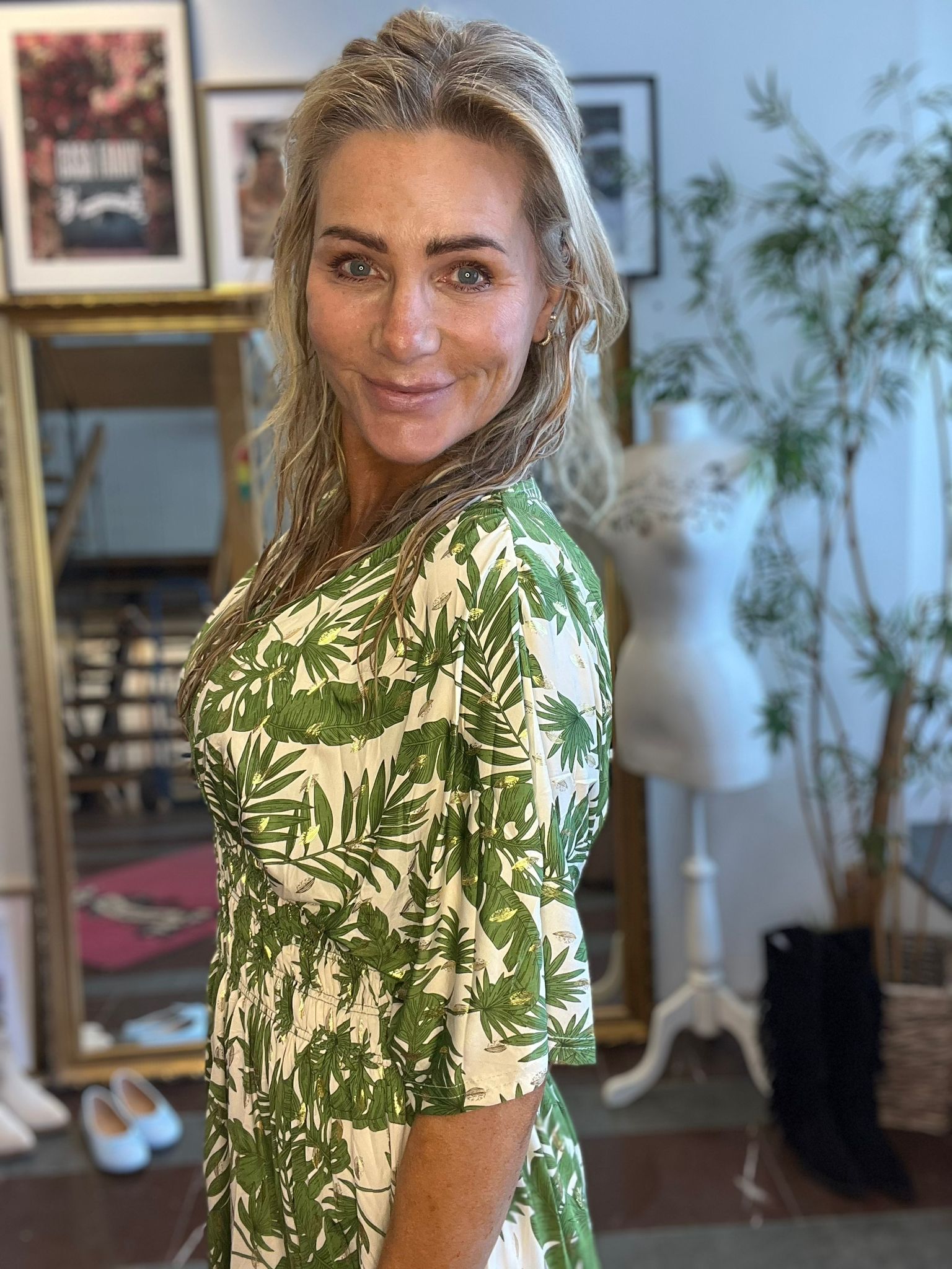 Leende kvinna med blont, vått hår vänder sig mot kameran och poserar i en vit klänning med grönt bladmönster. Hon står inomhus i ett rum med inramade tavlor, klädställning och stor krukväxt i bakgrunden.
