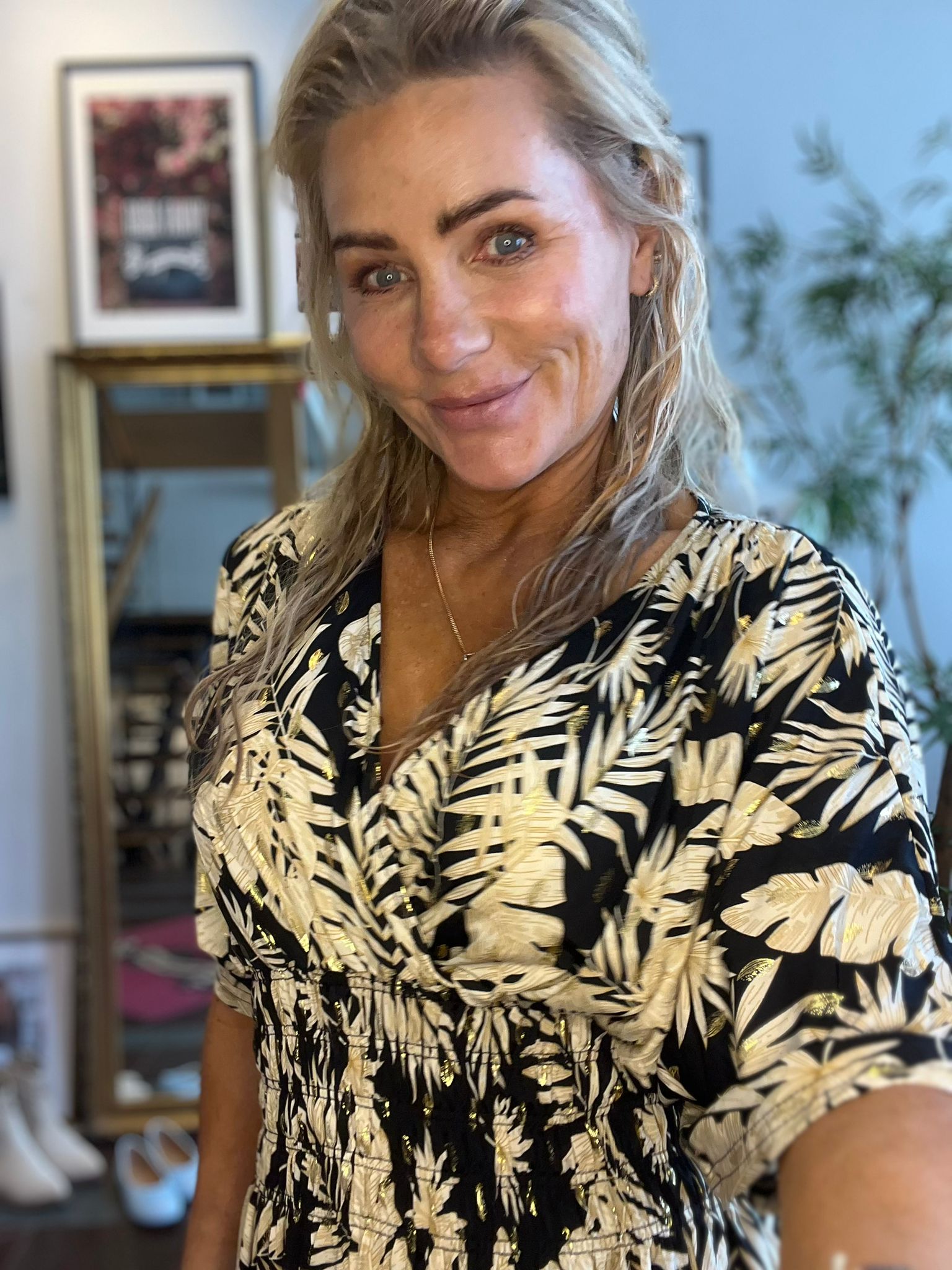 Leende person med långt blont hår tar en selfie inomhus, klädd i svart klänning med beige bladmönster och gulddetaljer. I bakgrunden syns en suddig tavla på väggen, en växt och möbler.