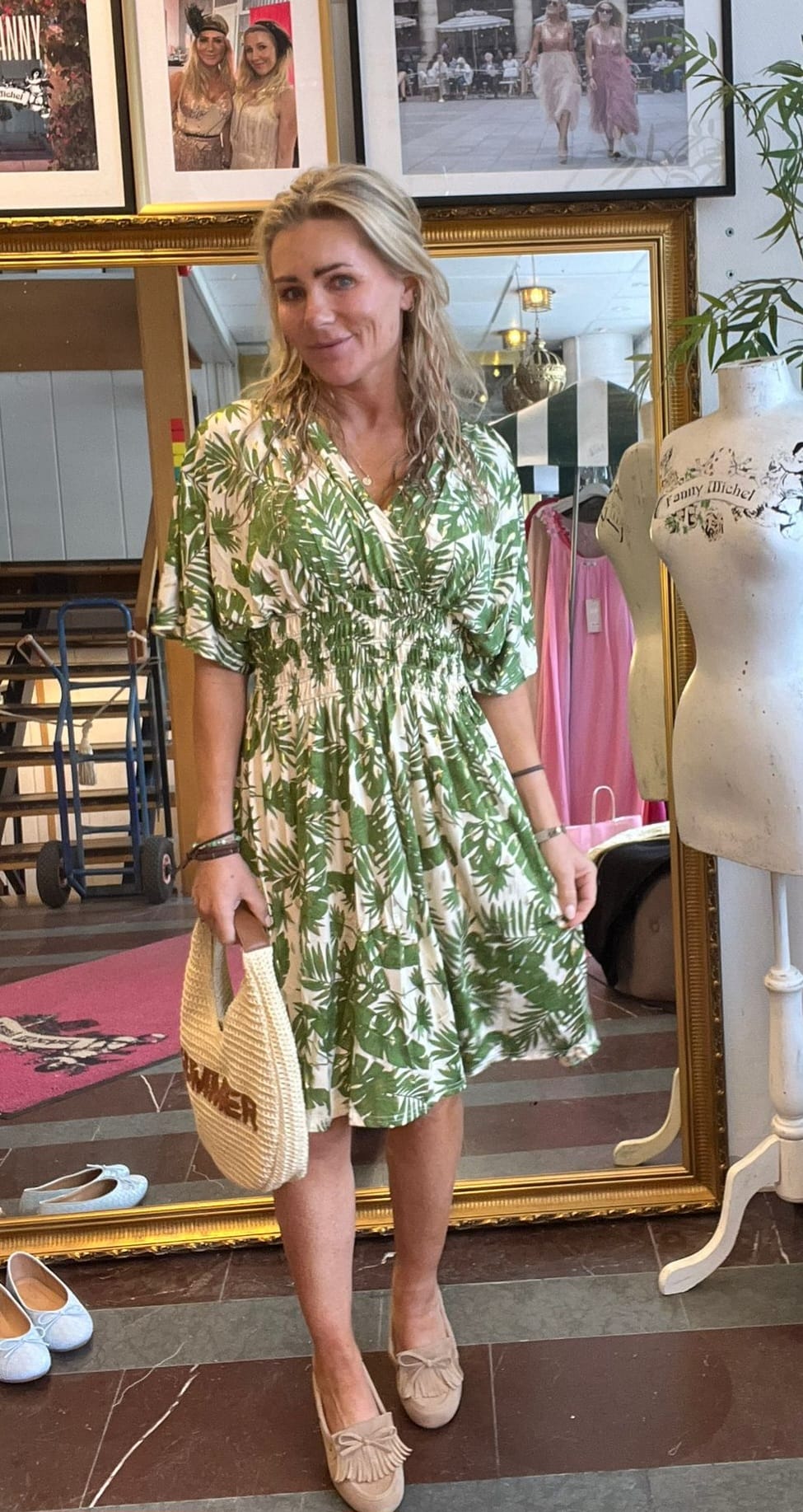 Kvinna står och posar i en butiksmiljö, klädd i grönvit bladmönstrad klänning och ljusa loafers, med en stråväska i handen. Bakom syns en stor spegel, modefoton och en provdocka med texten “Fanny Michel”.