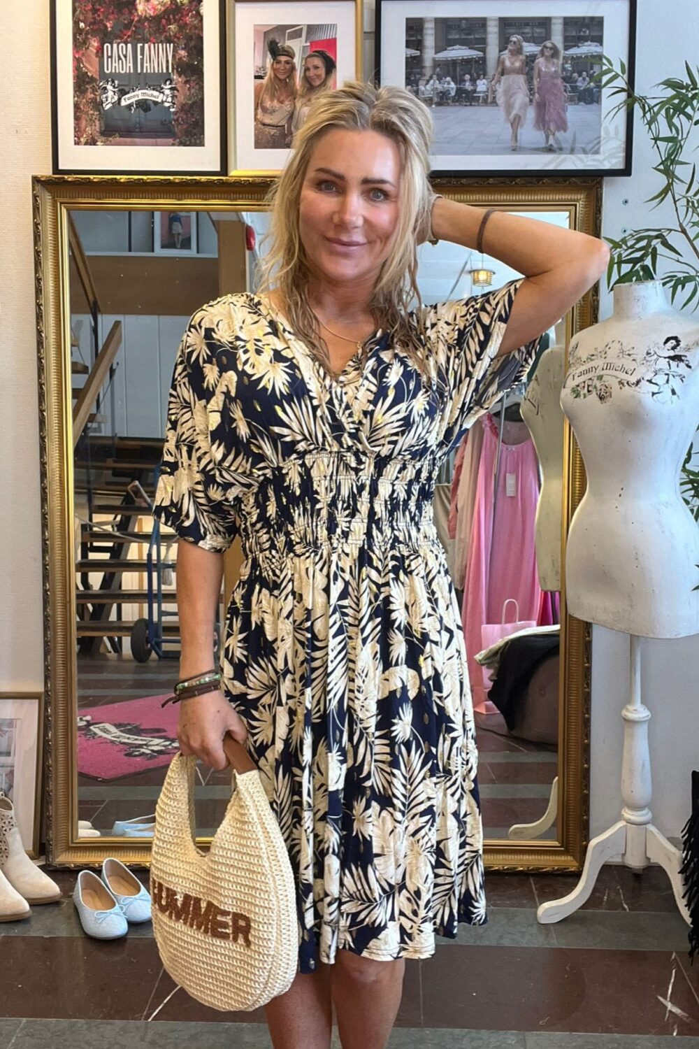 Blond kvinna står och ler i en klädbutik, med ena handen bakom huvudet och en stråväska där det står “SUMMER”. Bakom syns stor spegel, provdocka och inramade bilder med texten “CASA FANNY” och “Fanny Michel”.
