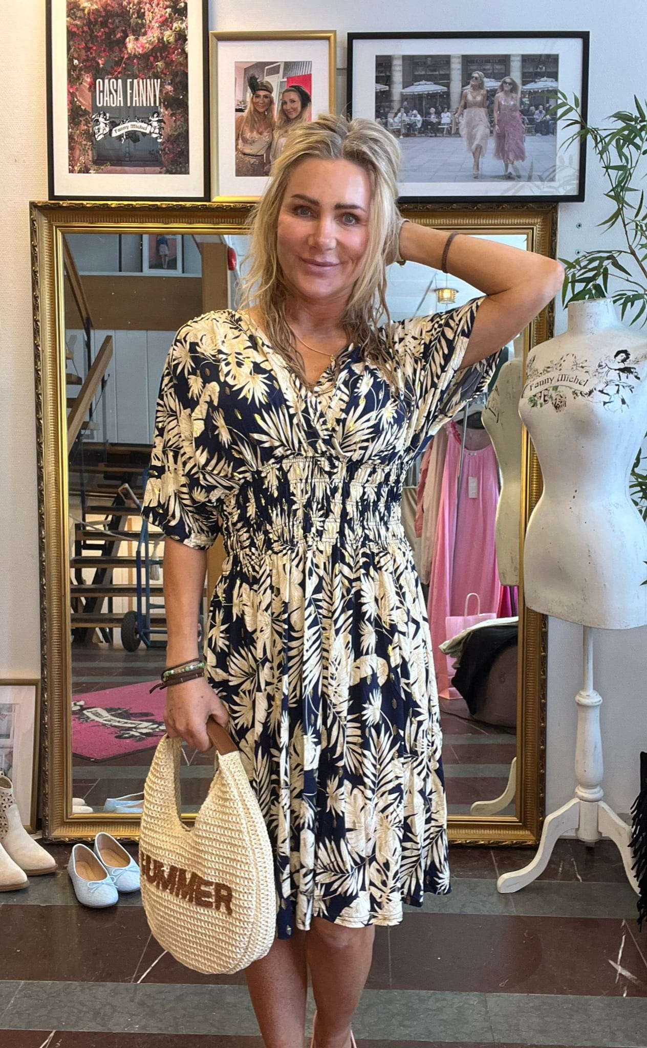 Blond kvinna står och ler i en klädbutik, med ena handen bakom huvudet och en stråväska där det står “SUMMER”. Bakom syns stor spegel, provdocka och inramade bilder med texten “CASA FANNY” och “Fanny Michel”.