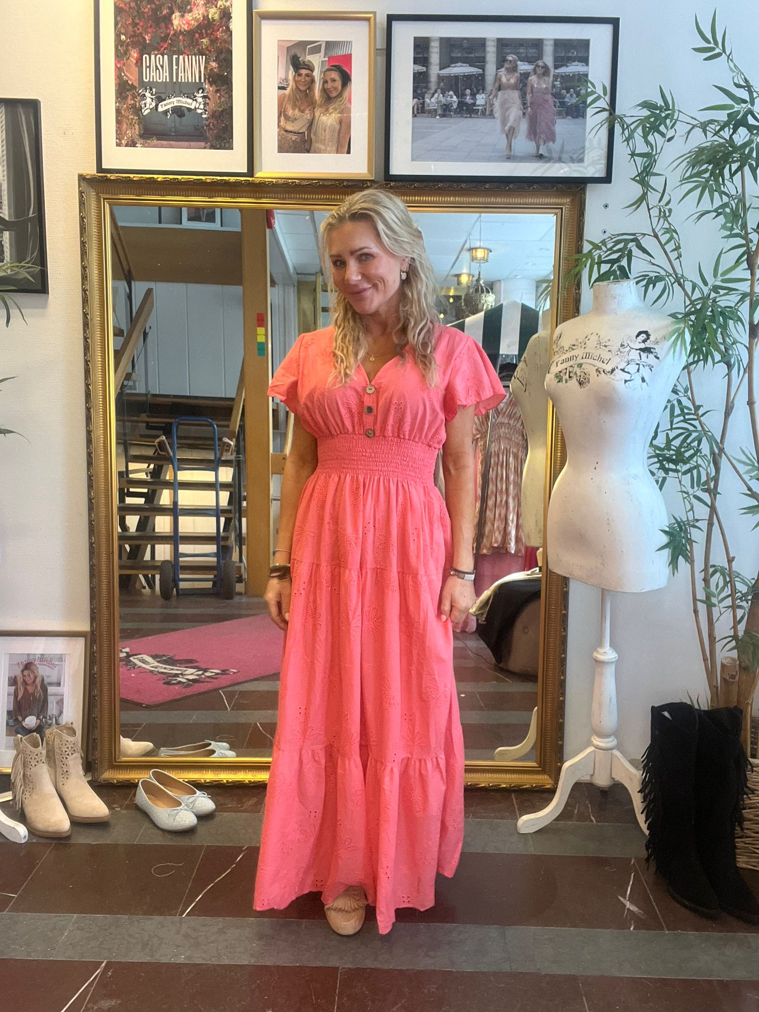 Kvinna i lång korallrosa klänning står och ler framför en stor guldramsspegel i en butik. Runt henne syns skor på golvet, provdocka med texten “Fanny Michel” och inramade foton, varav ett säger “Casa Fanny”.