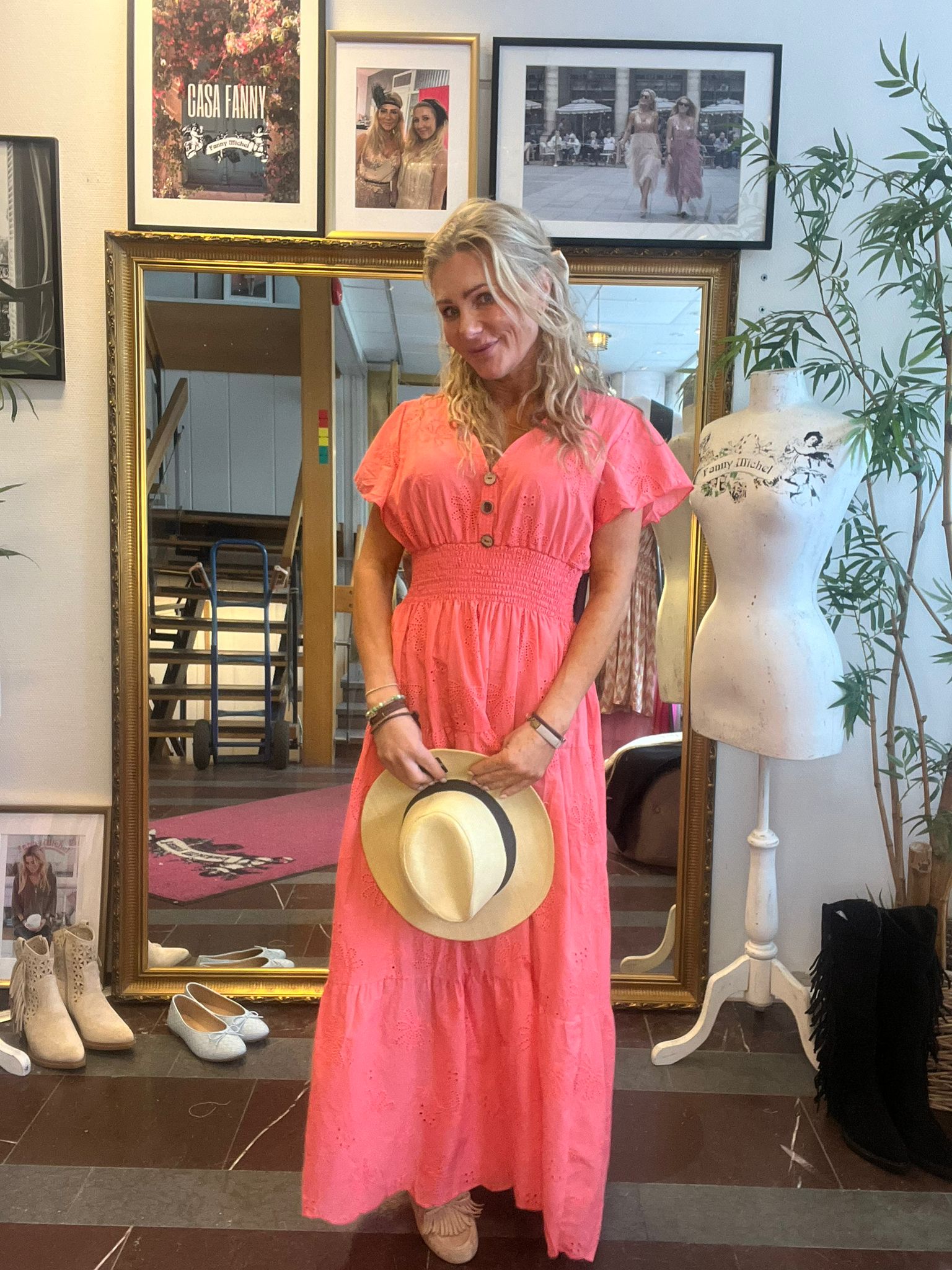 Kvinna i korallrosa långklänning står och ler, håller en ljus stråhatt framför sig i en butik eller ateljé. Bakom henne syns stor guldramsspegel, skor på golvet, provdocka och inramade foton med texten “CASA FANNY”.