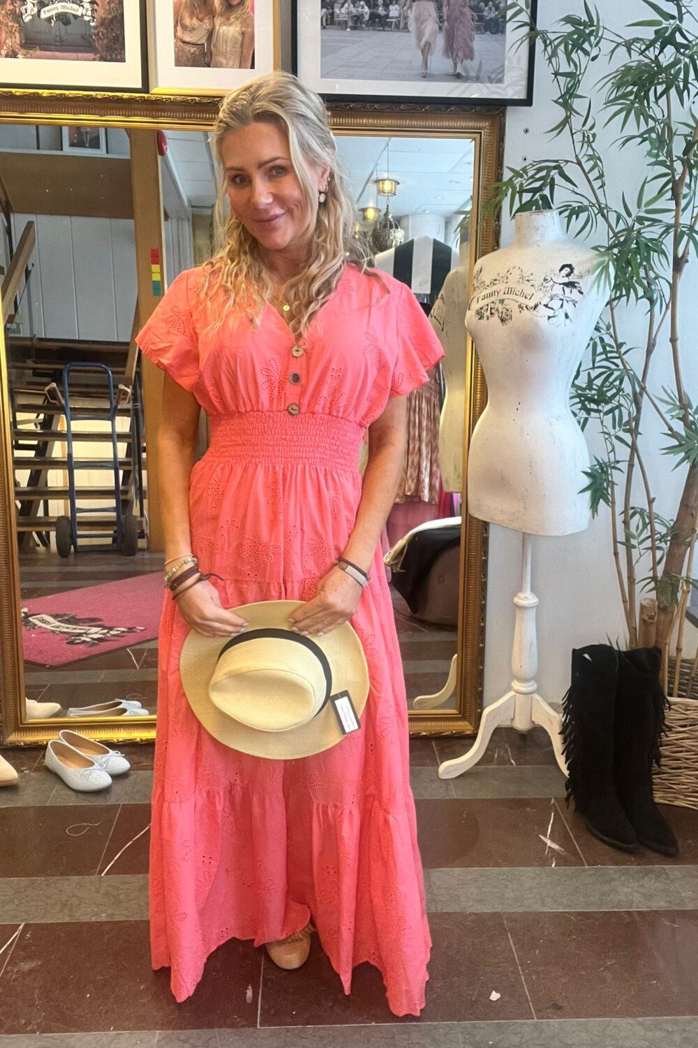 Kvinna i korallrosa långklänning står och ler, håller en stråhatt framför sig i en butik. Bakom henne syns en stor guldramsspegel, skor på golvet, provdocka med texten “Fanny Michel” och en grön växt.