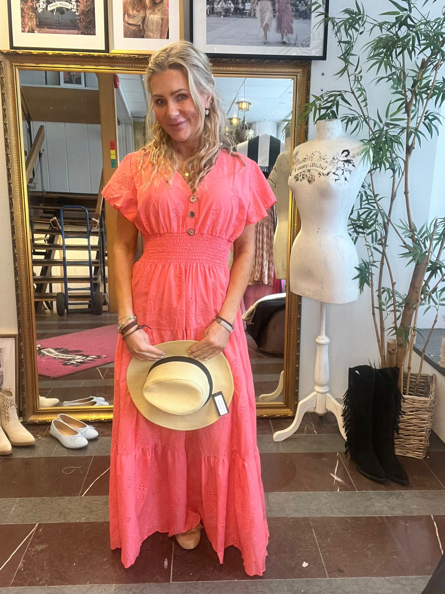Kvinna i korallrosa långklänning står och ler, håller en stråhatt framför sig i en butik. Bakom henne syns en stor guldramsspegel, skor på golvet, provdocka med texten “Fanny Michel” och en grön växt.