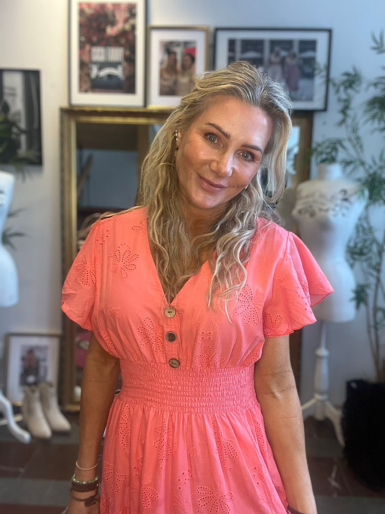 Leende kvinna med långt blont hår lutar huvudet lätt och tittar mot kameran, klädd i en korallrosa klänning med broderade blommor och knappar. Hon står i ett hemrum med spegel, tavlor och växter i bakgrunden.