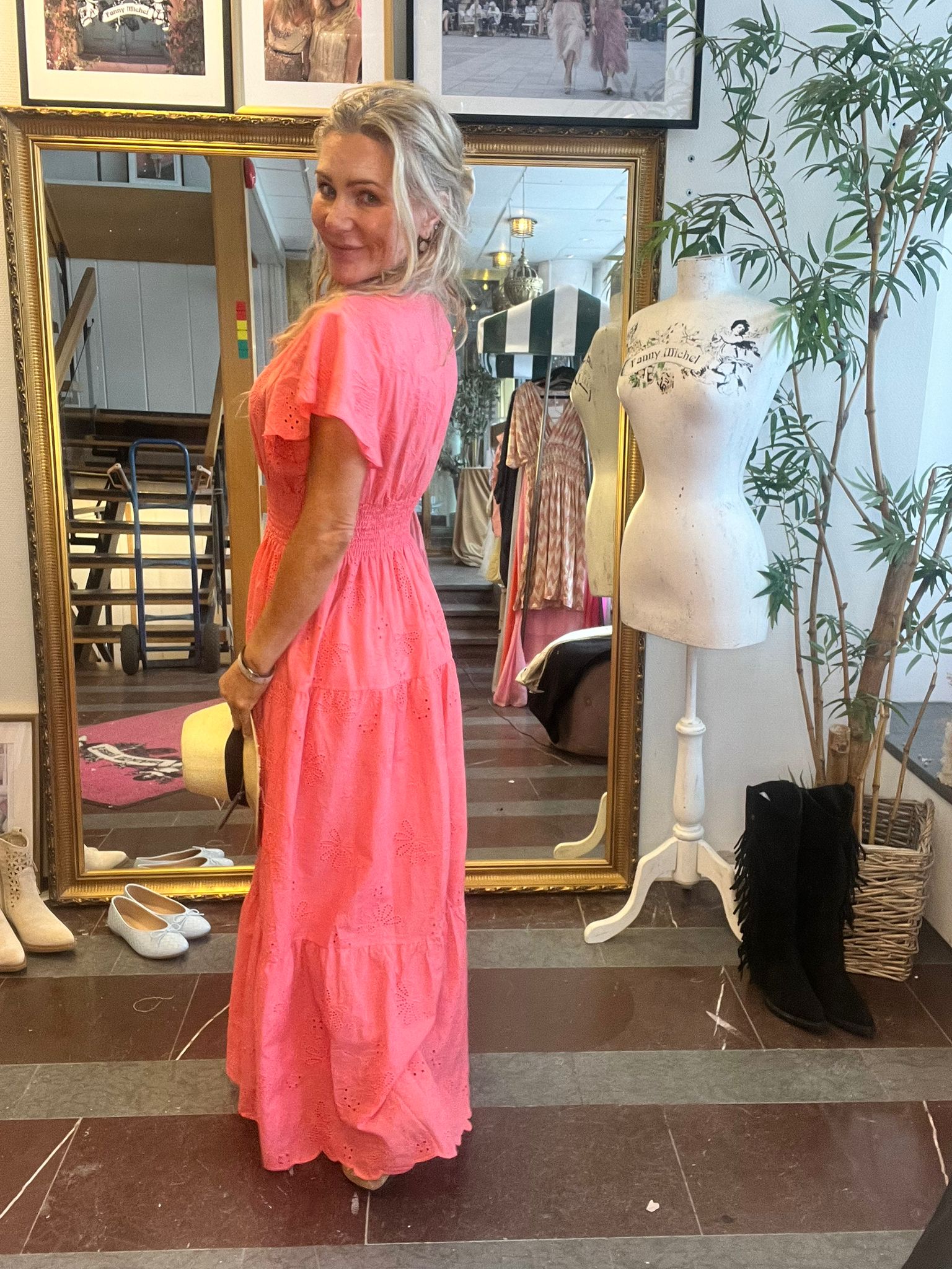 Kvinna i lång korallrosa klänning står och ler med kroppen vänd åt sidan, håller en hatt och tittar mot kameran. Hon är framför en stor guldramsspegel i en butik, med skyltdocka märkt “Fanny Michel”.