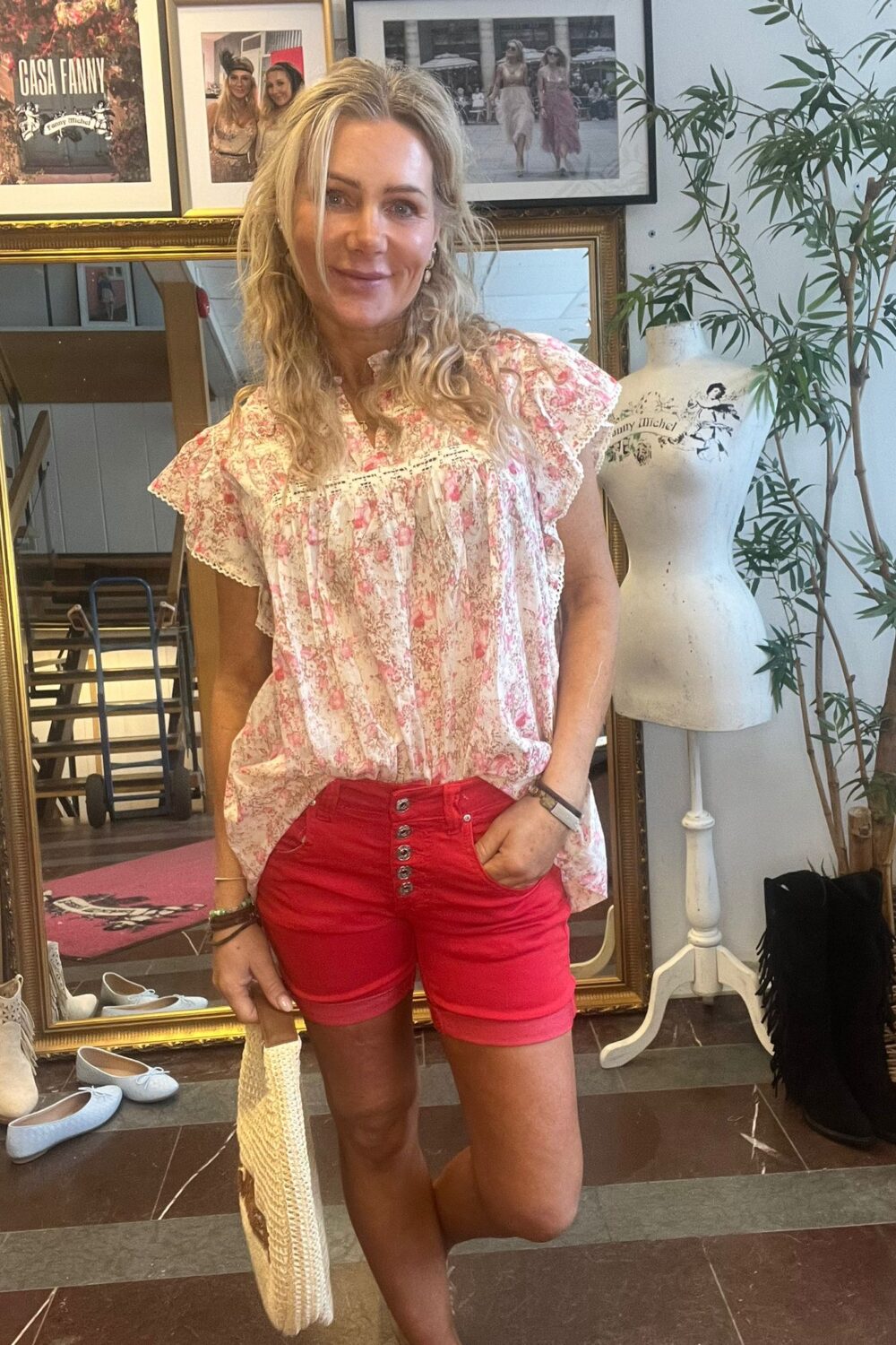 Leende kvinna står med handen i fickan och håller en vävd väska i en butik eller ateljé. Hon bär blommig topp och röda shorts; bakom syns spegel, skyltdocka, skor och inramade foton med texten ”GASA FANNY”.