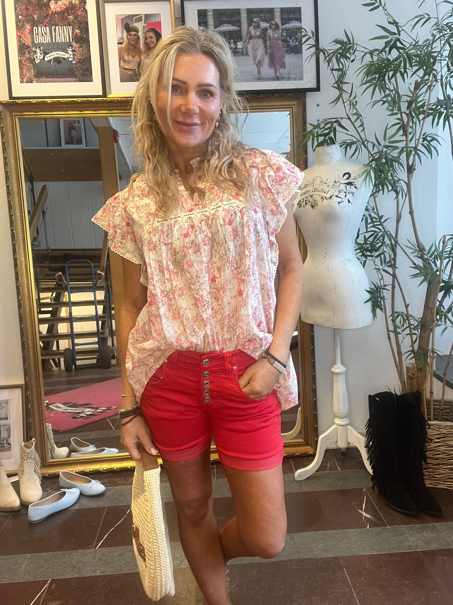 Leende kvinna står med handen i fickan och håller en vävd väska i en butik eller ateljé. Hon bär blommig topp och röda shorts; bakom syns spegel, skyltdocka, skor och inramade foton med texten ”GASA FANNY”.