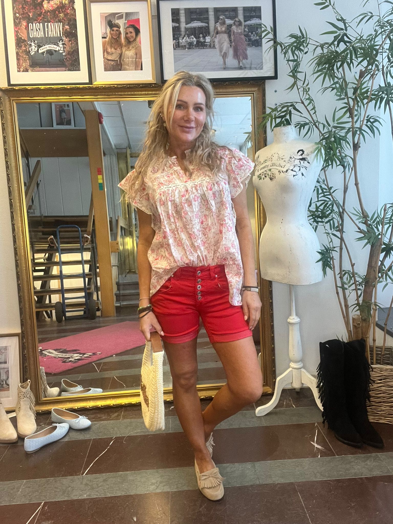Leende kvinna poserar med en halmväska framför en stor guldramsspegel i en butik/ateljé, klädd i blommig topp och röda shorts. På väggen syns tavlor med texten ”CASA FANNY” och en provdocka märkt ”Fanny Michel”.
