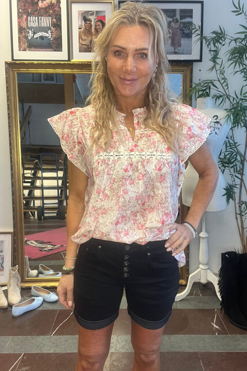 Blond kvinna står och ler med handen på höften, klädd i rosa blommig blus och svarta shorts, i en inredningsmiljö med spegel, skor och växt. Inramad affisch visar texten “CASH FANNY”.