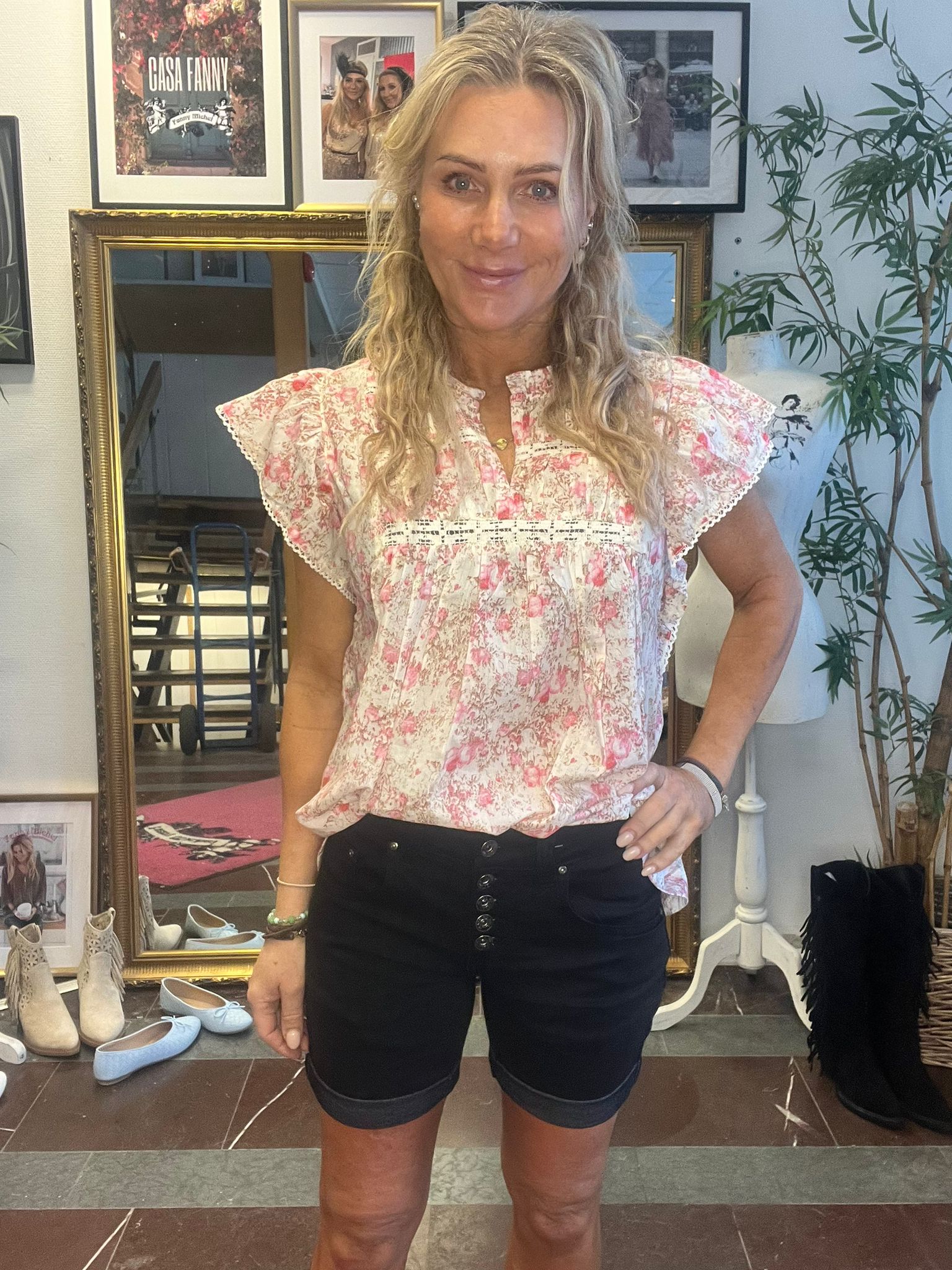 Blond kvinna står och ler med handen på höften, klädd i rosa blommig blus och svarta shorts, i en inredningsmiljö med spegel, skor och växt. Inramad affisch visar texten “CASH FANNY”.