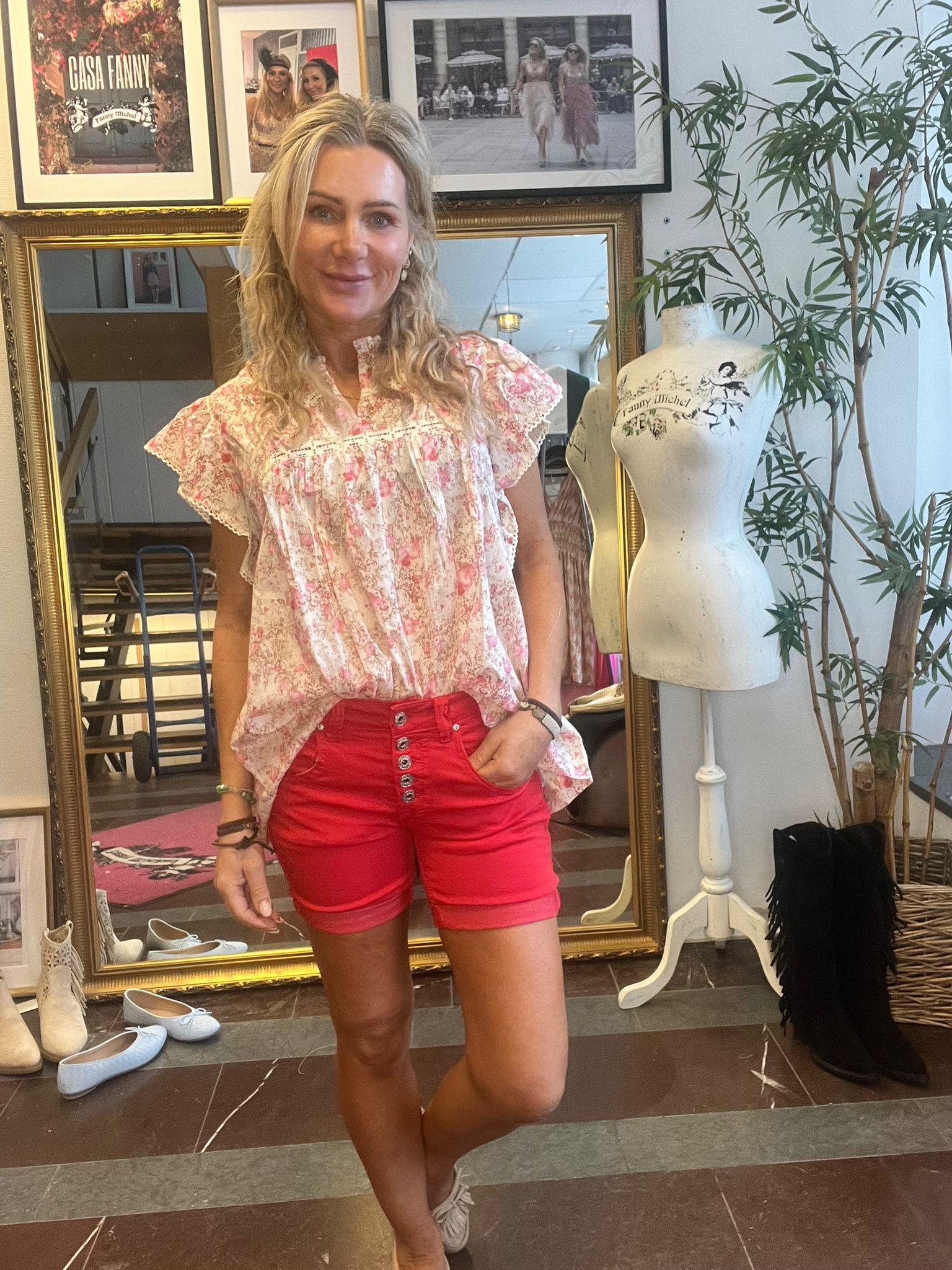 Kvinna står och ler med ena handen i fickan, klädd i blommig rosa blus och röda shorts. Hon poserar framför en guldramsspegel i en butik med provdocka, skor på golvet och tavlor med texten “CASH FANNY” och “Fanny Michel”.