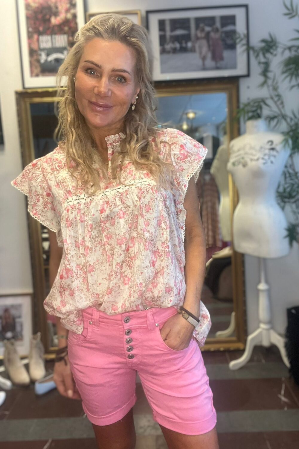 Leende kvinna står och poserar med ena handen i fickan, klädd i vit blommig volangtopp och rosa shorts. Bakom henne syns en klädbutik/ateljé med inramade foton på väggen och en provdocka.